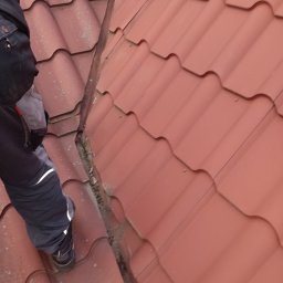 Dekarstwo Blacharstwo Sebastian Karliński JDG - Fragment dachu z czerwonej dachówki ceramicznej, widoczne zabrudzenia i mech w szczelinach, obok stoi dekarz w roboczym ubraniu i rękawicach.