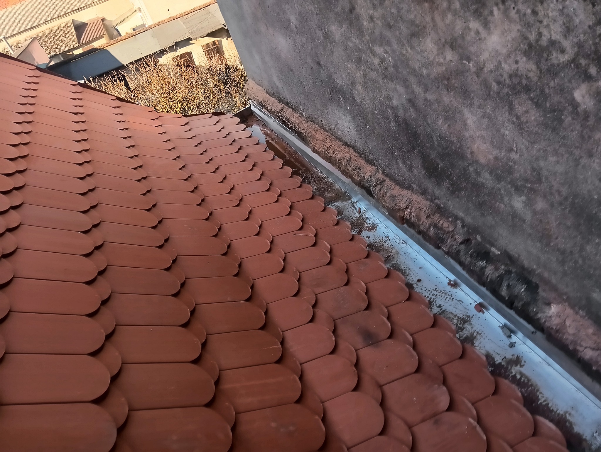 Nowe, brązowe dachówki ułożone na dachu, widok z góry, obok ściana budynku i rynna.