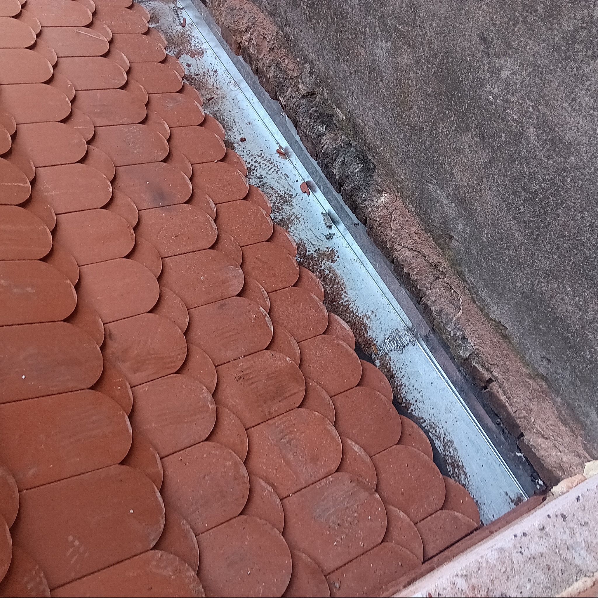 Widok z góry na fragment dachu pokrytego czerwoną dachówką karpiówką, z widoczną, nową metalową rynną wzdłuż krawędzi dachu przylegającej do ściany.