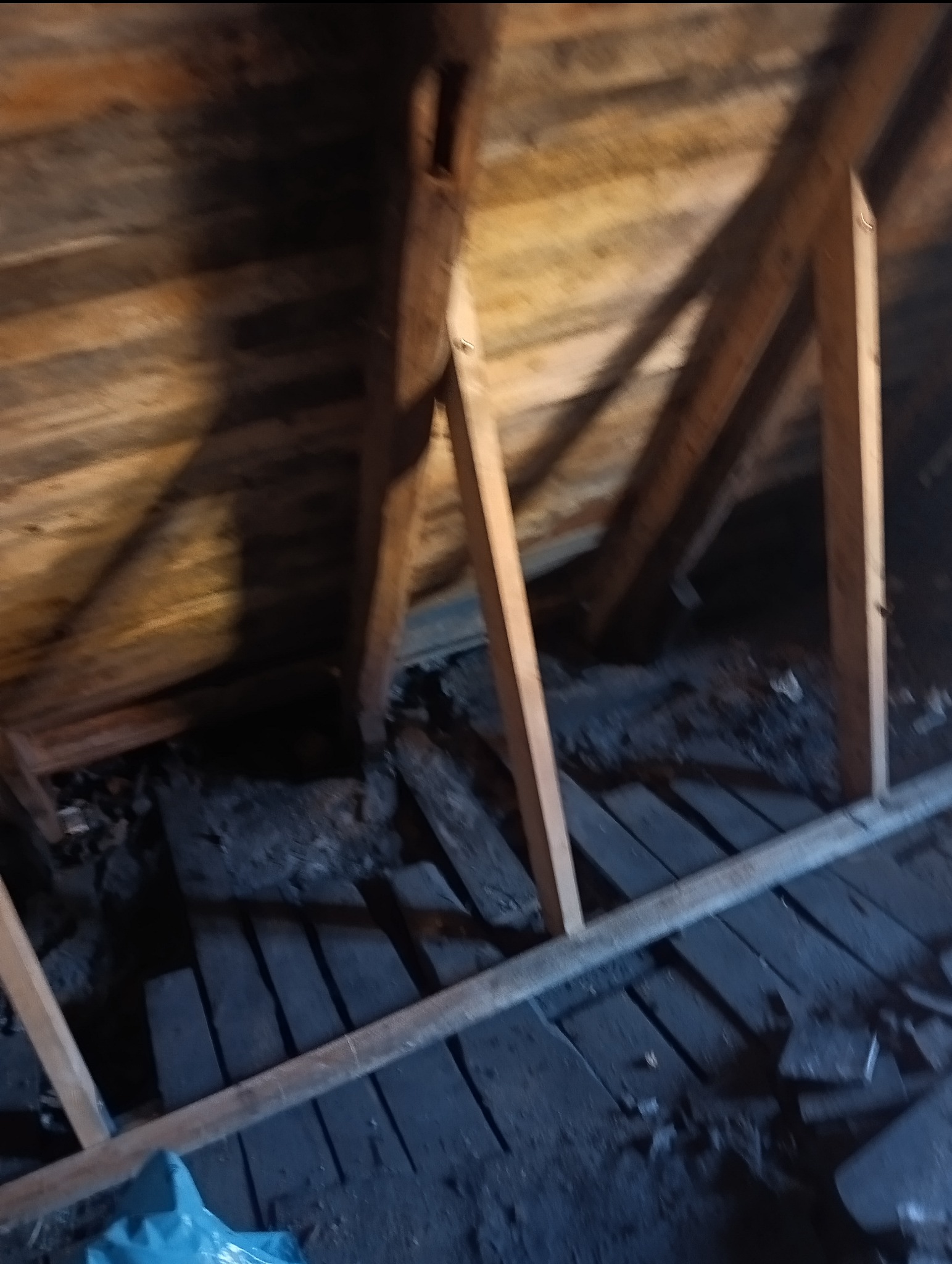 Wnętrze poddasza z widoczną drewnianą konstrukcją więźby dachowej, deskami poszycia i elementami starej izolacji. Widoczne fragmenty podłogi. Niska jakość zdjęcia.