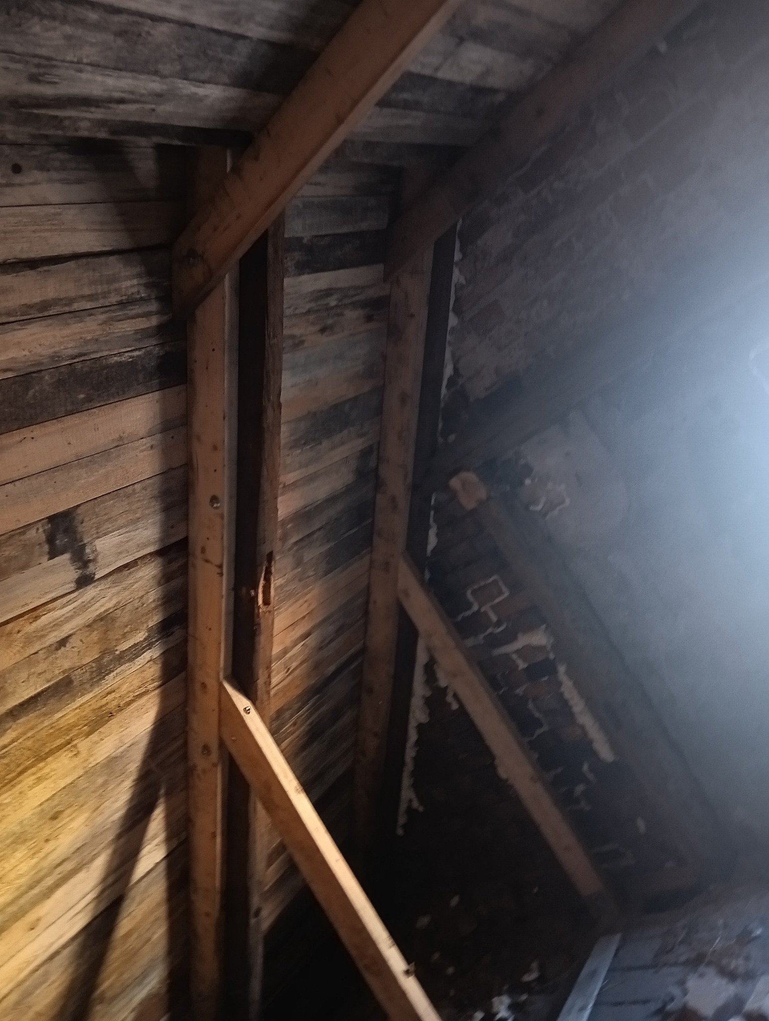 Surowe drewniane elementy konstrukcyjne poddasza, widoczne fragmenty starej cegły i desek, słabe oświetlenie.