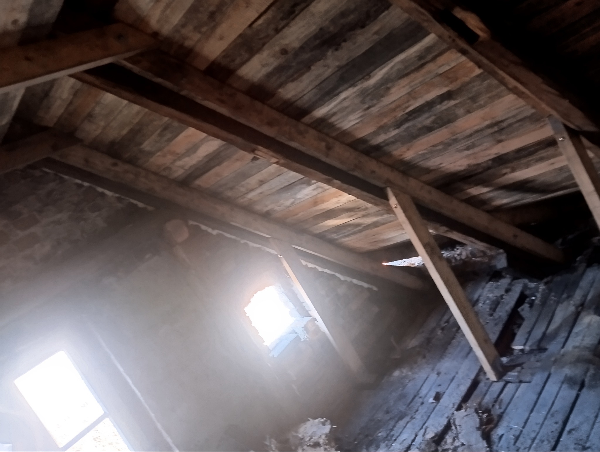 Wnętrze poddasza z widoczną drewnianą konstrukcją dachu, deski z widocznymi śladami zużycia, w tle okno z silnym światłem dziennym.