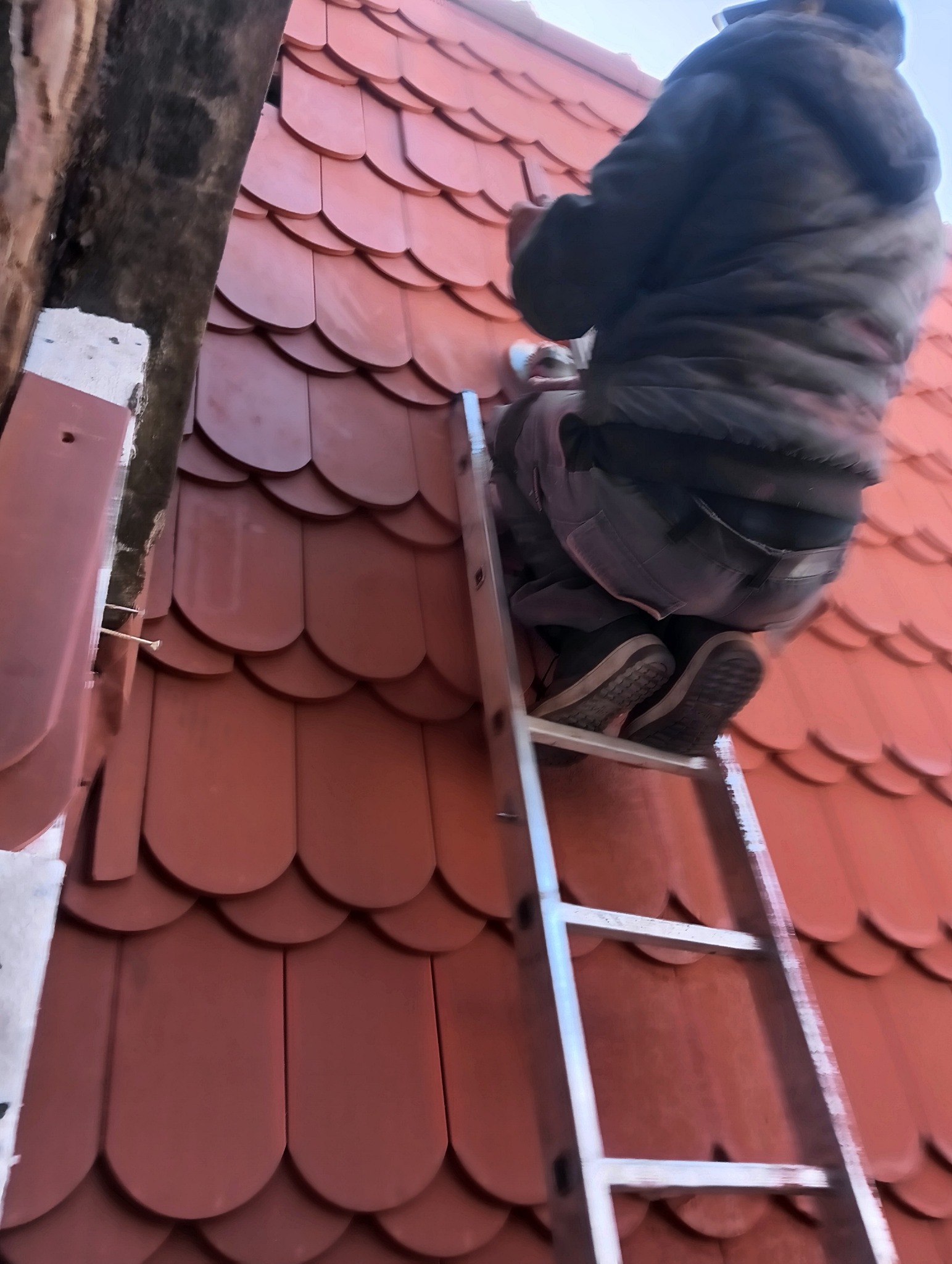 Pracownik na drabinie, montujący dachówkę karpiówkę w kolorze ceglastym, widok z dołu na fragment dachu i osobę w roboczym ubraniu.
