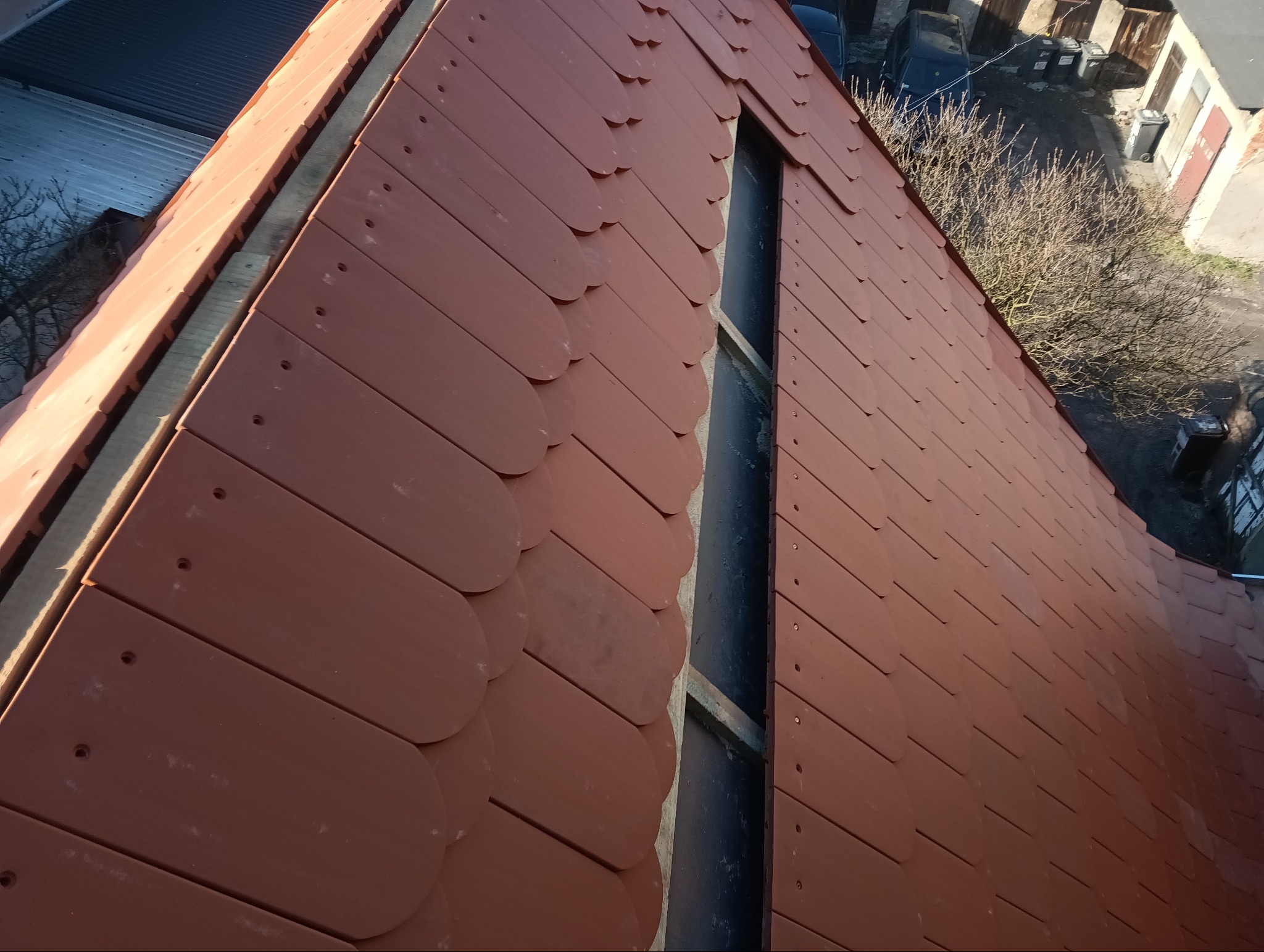 Nowe, brązowe dachówki ceramiczne w kształcie łuski, ułożone na połaci dachu z oknem połaciowym, widok z góry na budynki gospodarcze w tle.