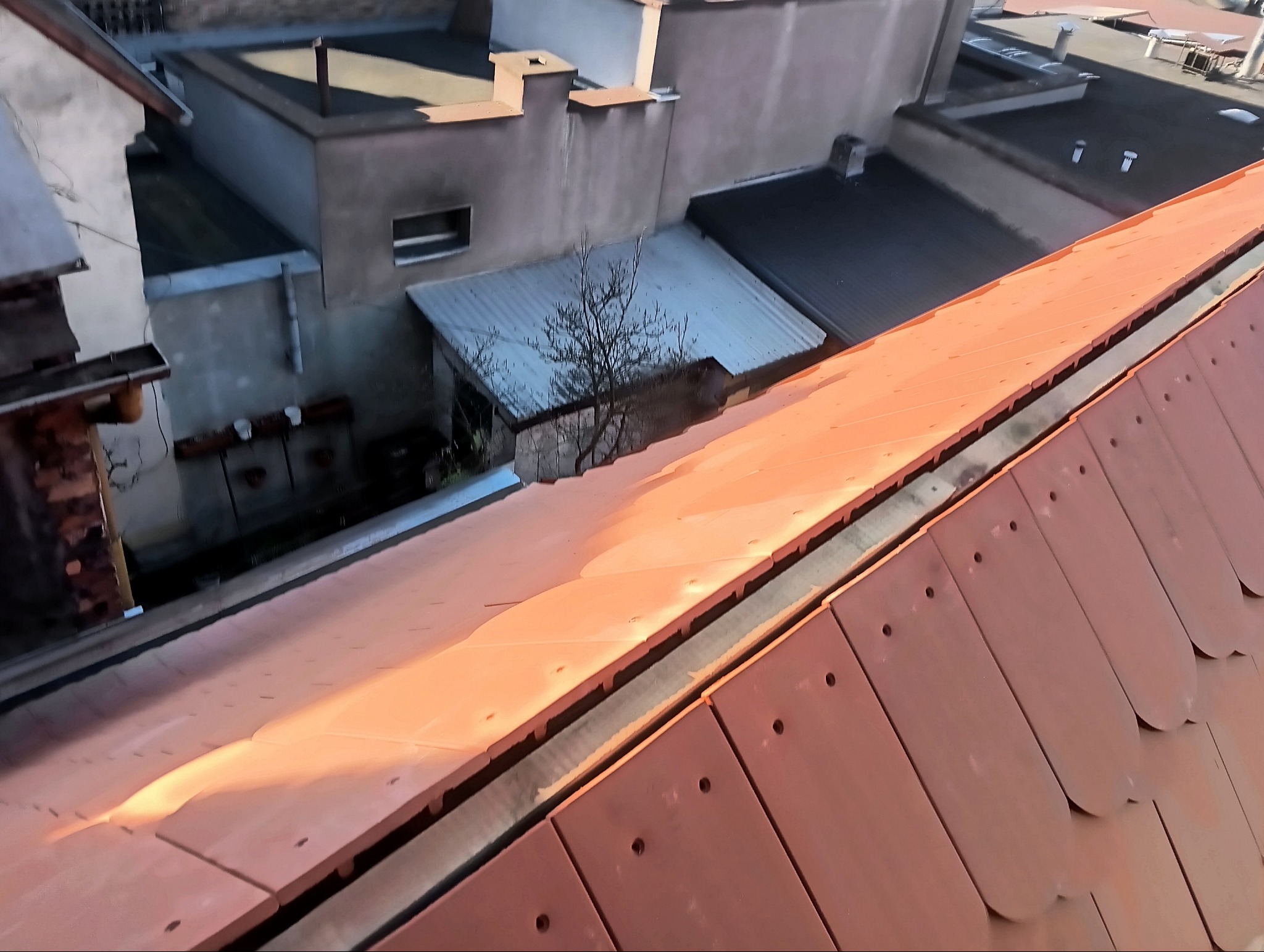 Nowy dach z dachówki ceramicznej w kolorze ceglastym, widok z góry na fragment dachu z widoczną konstrukcją i sąsiednimi budynkami w tle.