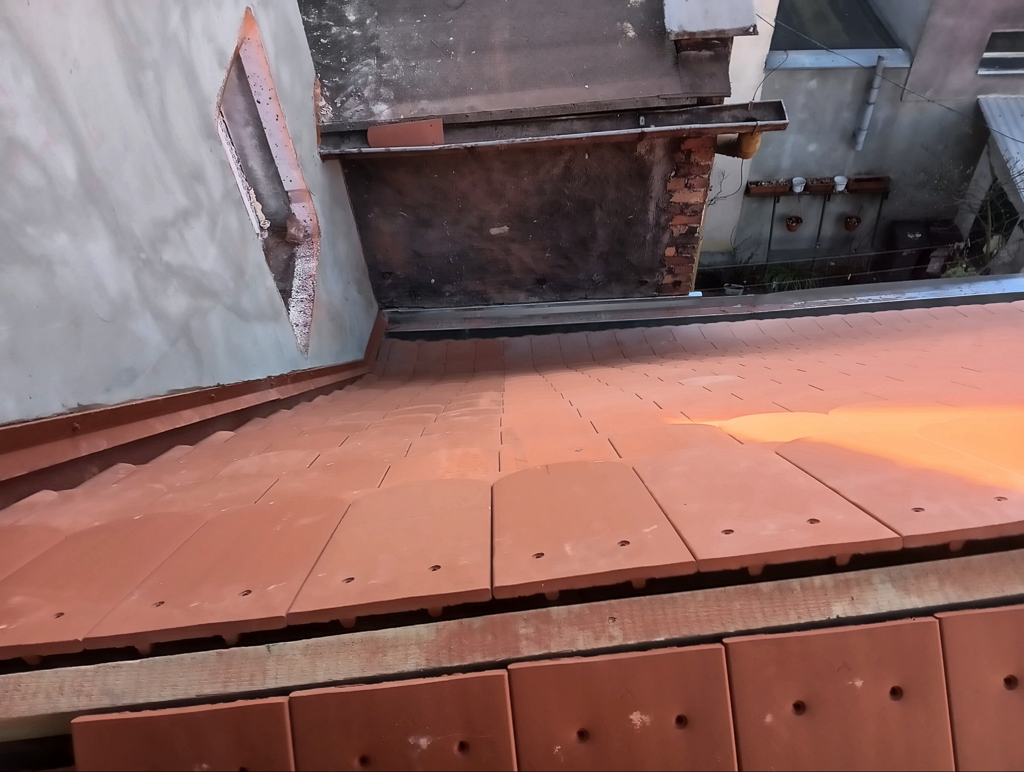 Widok z góry na częściowo ułożone, nowe, terakotowe dachówki na drewnianej konstrukcji, z fragmentem ściany budynku z otworem w tle.