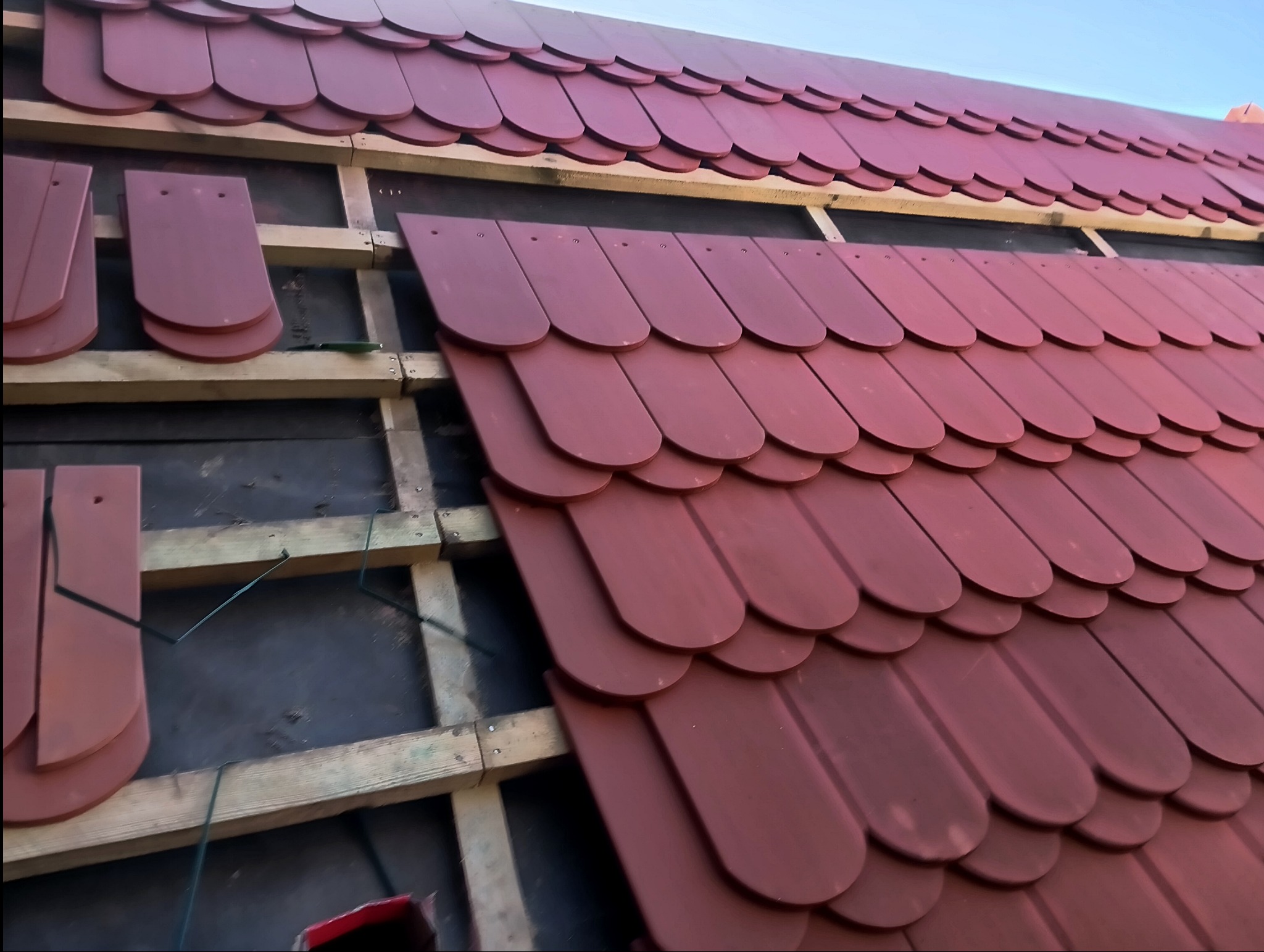 Montaż bordowej dachówki karpiówki na drewnianej konstrukcji dachu, widoczne fragmenty membrany dachowej i niezamontowane płytki.