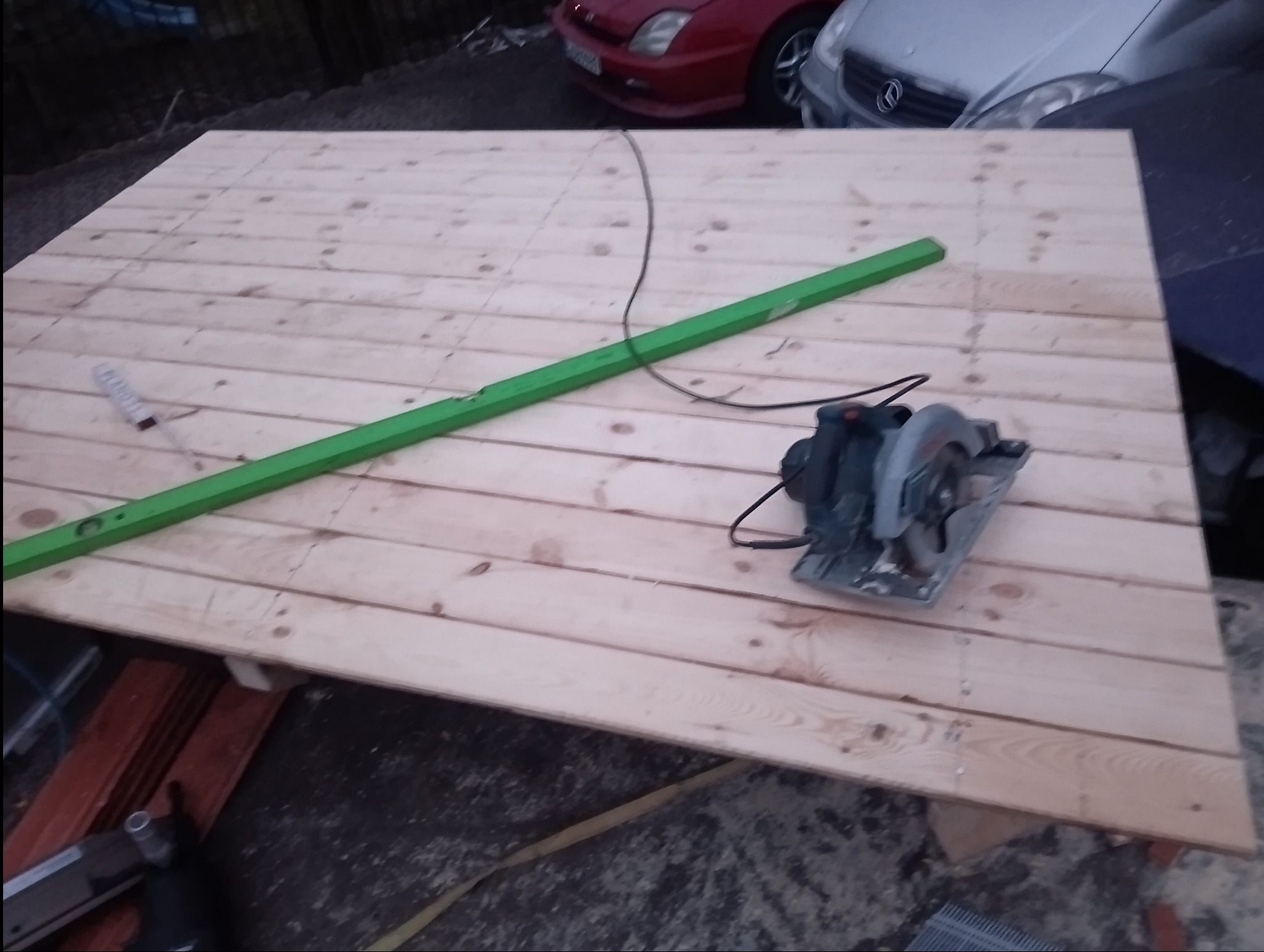 Przygotowanie drewnianej konstrukcji dachu: deski ułożone na legarach, poziomica i piła elektryczna w kadrze, samochody w tle.