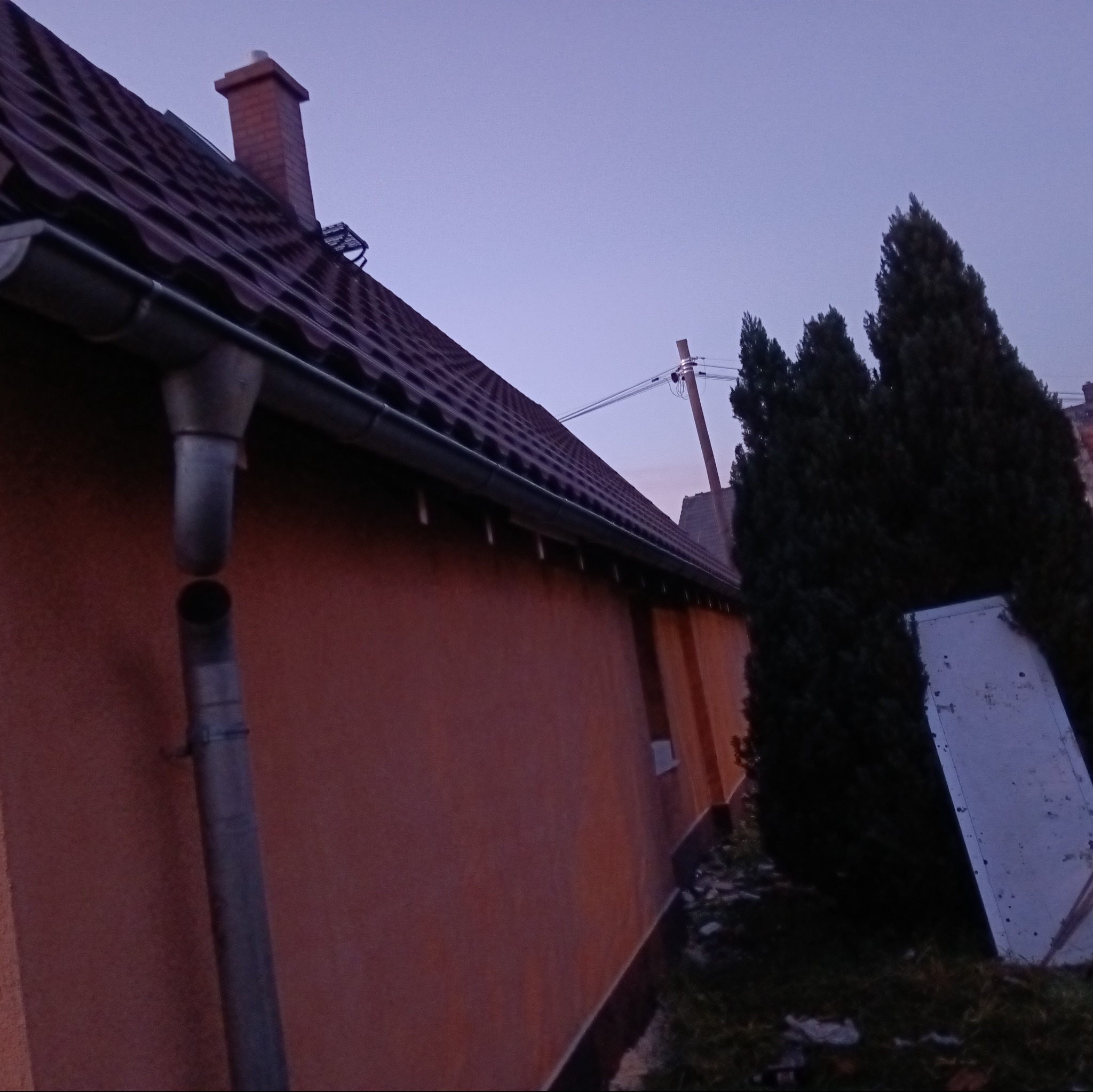 Czerwono-brązowy dach z kominem i rynną, widok z dołu na tle wieczornego nieba i drzew