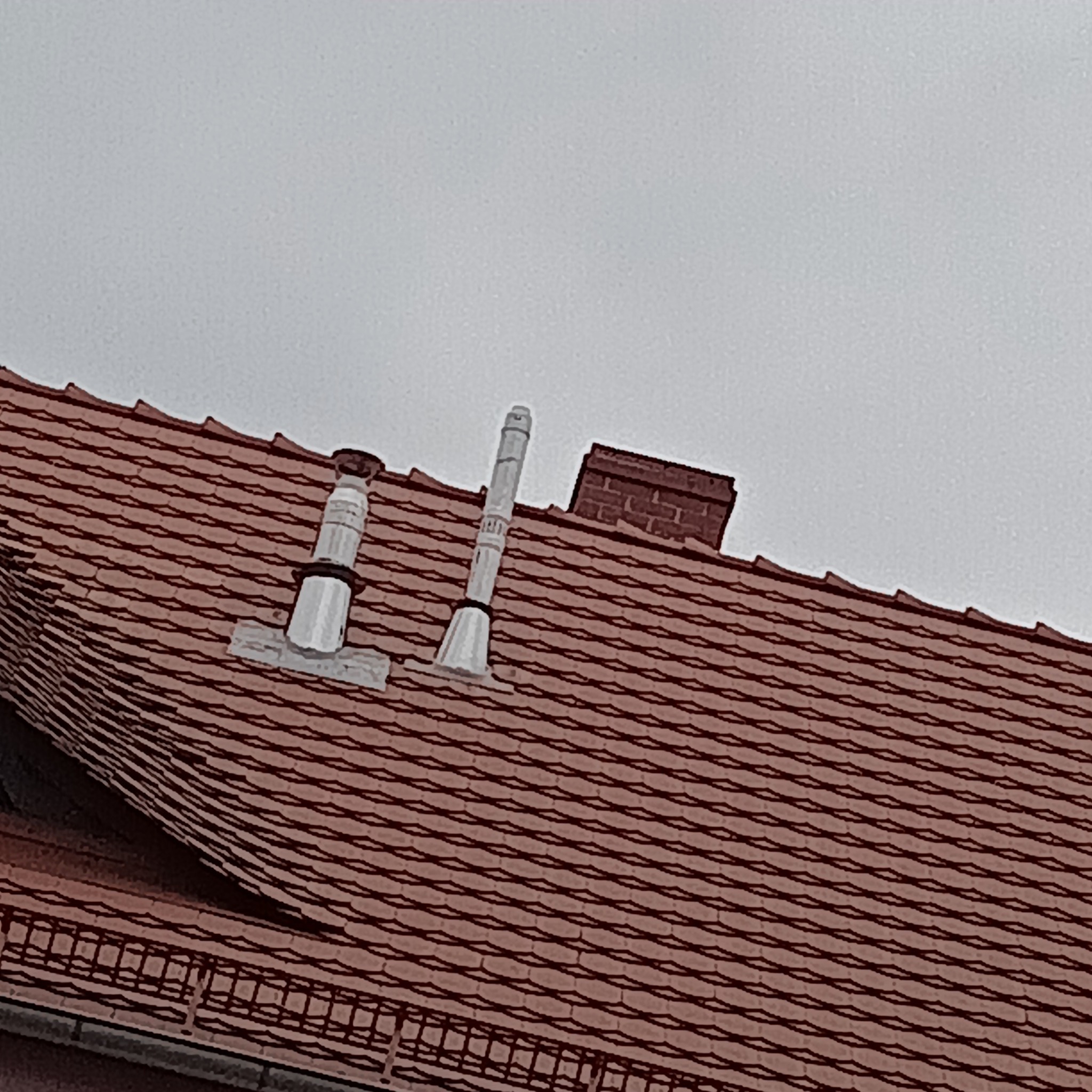 Czerwona dachówka ceramiczna z dwoma kominami wentylacyjnymi i ceglanym kominem w tle, widok z góry.