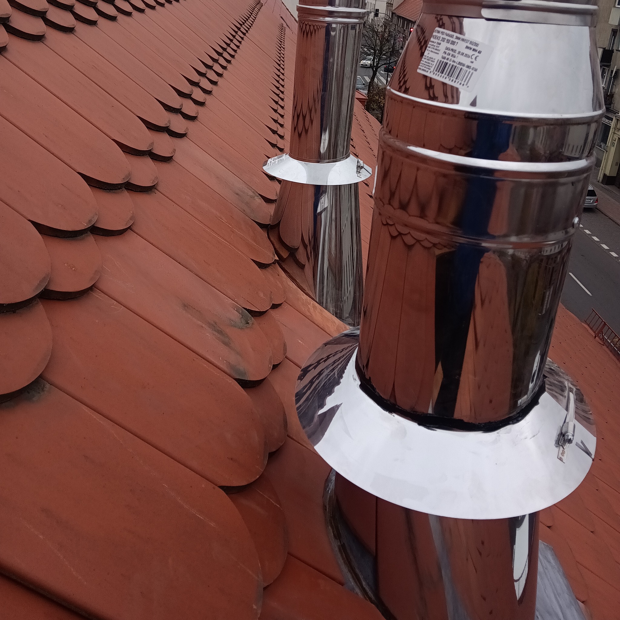 Dwa nowe, błyszczące kominy ze stali nierdzewnej wystają z dachu pokrytego czerwoną dachówką karpiówką, widok z góry na tle miejskiej zabudowy.