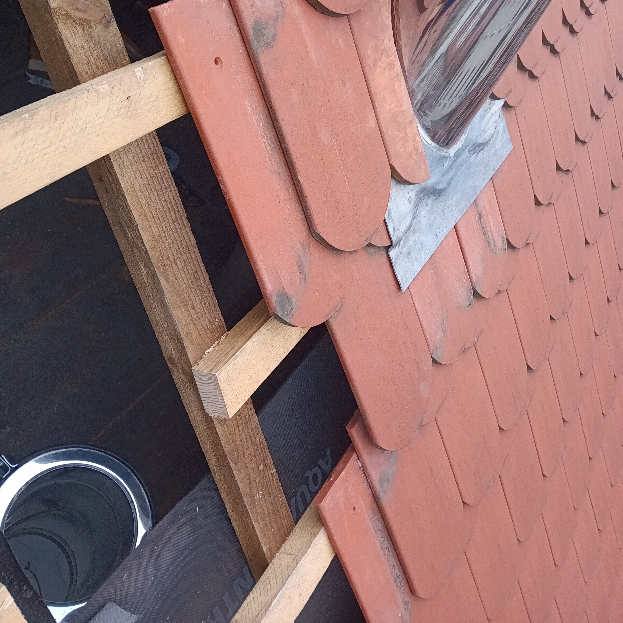 Fragment dachu z czerwoną dachówką karpiówką, widoczna więźba dachowa i obróbka blacharska komina, fragment membrany dachowej.