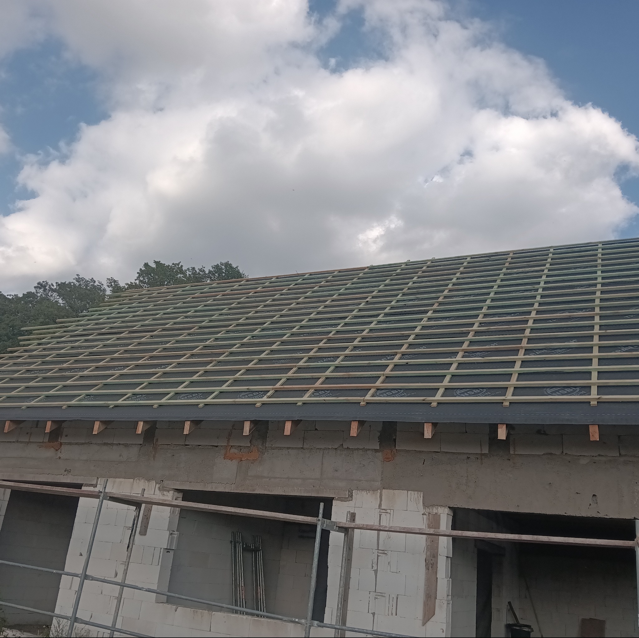 Konstrukcja dachu z łatami drewnianymi na czarnej membranie, widok z dołu na tle błękitnego nieba z chmurami, budynek w trakcie budowy z widocznym rusztowaniem.