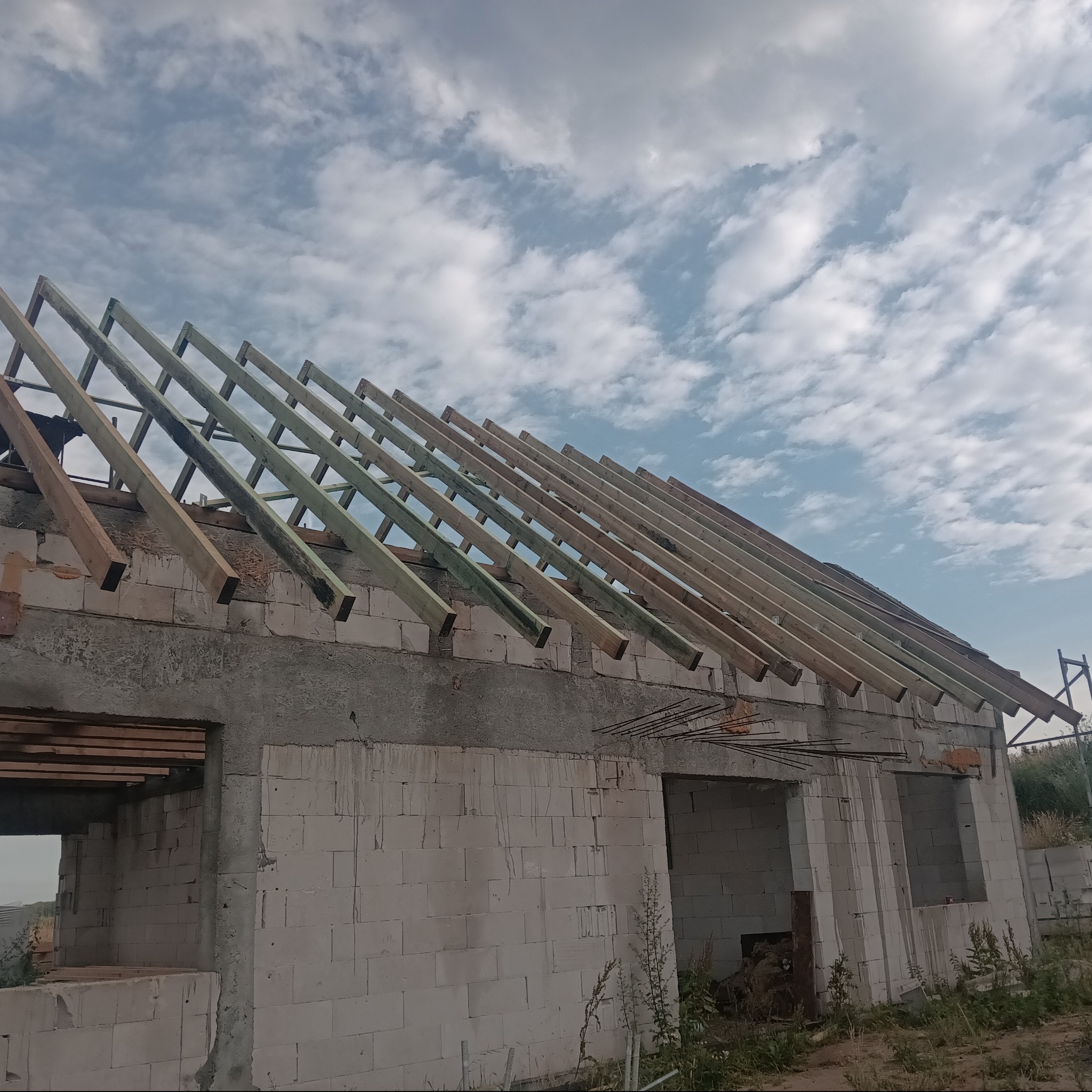 Wznoszenie więźby dachowej na surowym budynku z białych bloczków, widoczne elementy konstrukcyjne i niebo z chmurami.
