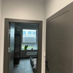electrician.on.site - Wnętrze mieszkania po remoncie: świeżo pomalowane ściany w odcieniach szarości, widoczne dwie nowoczesne, grafitowe ościeżnice i drzwi, w tle okno z parapetem i elementami wyposażenia pokoju.