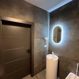electrician.on.site - Nowoczesna łazienka z ciemnymi płytkami, wolnostojącą umywalką oświetloną ledowym lustrem, oraz minimalistycznymi drzwiami z poziomymi frezami.