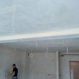 electrician.on.site - Mężczyzna szpachluje ścianę w pomieszczeniu w stanie deweloperskim, widoczne przewody elektryczne zwisające z sufitu i zabezpieczony klimatyzator.