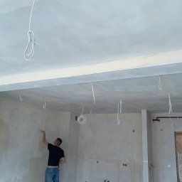 electrician.on.site - Mężczyzna w czarnej koszulce szpachluje ścianę w surowym wnętrzu z wystającymi przewodami elektrycznymi z sufitu, przygotowując pomieszczenie do malowania.