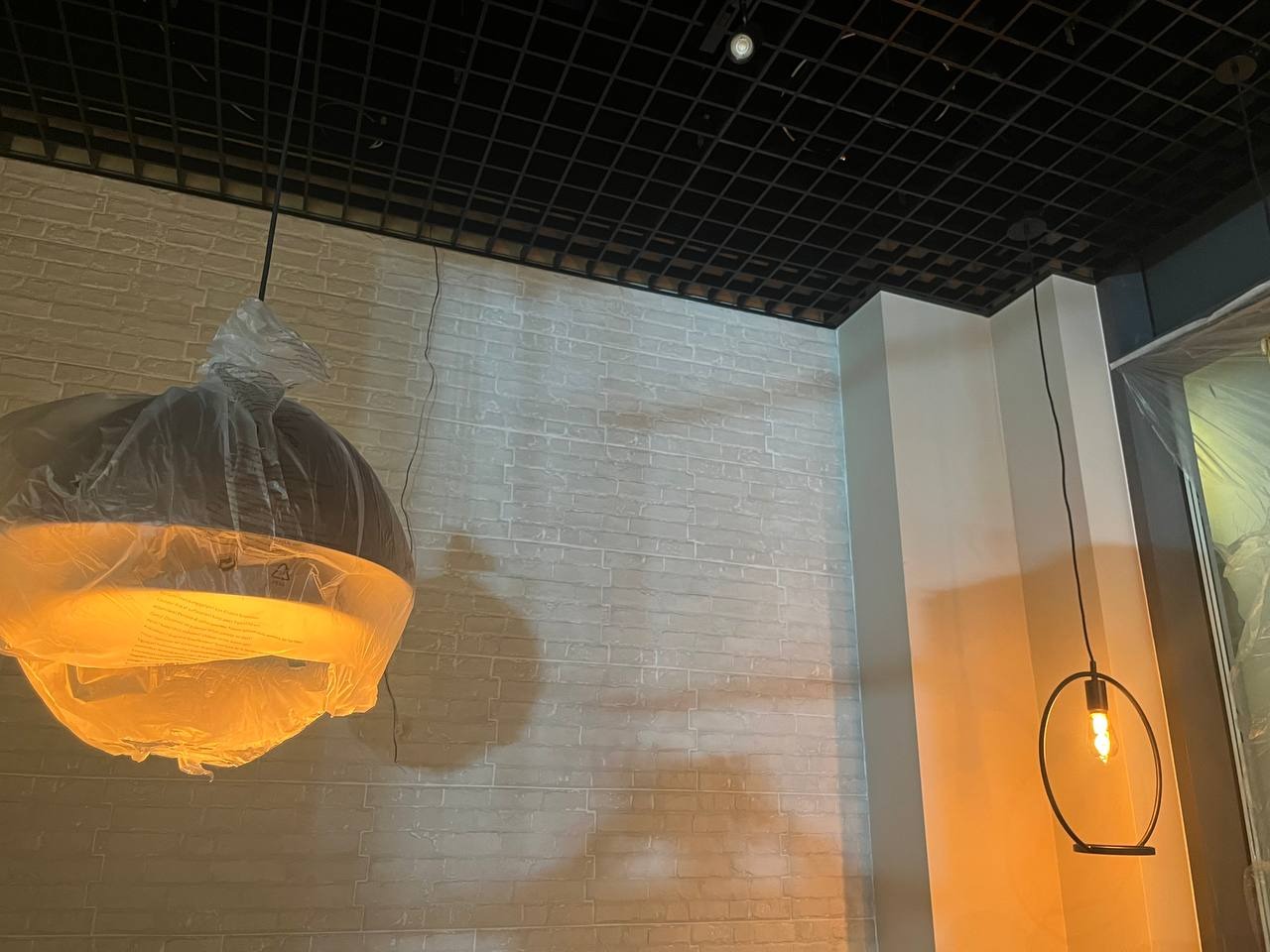 Nowe lampy wiszące: jedna okrągła w folii ochronnej, druga minimalistyczna z żarówką, na tle ściany z cegły i sufitu rastrowego.