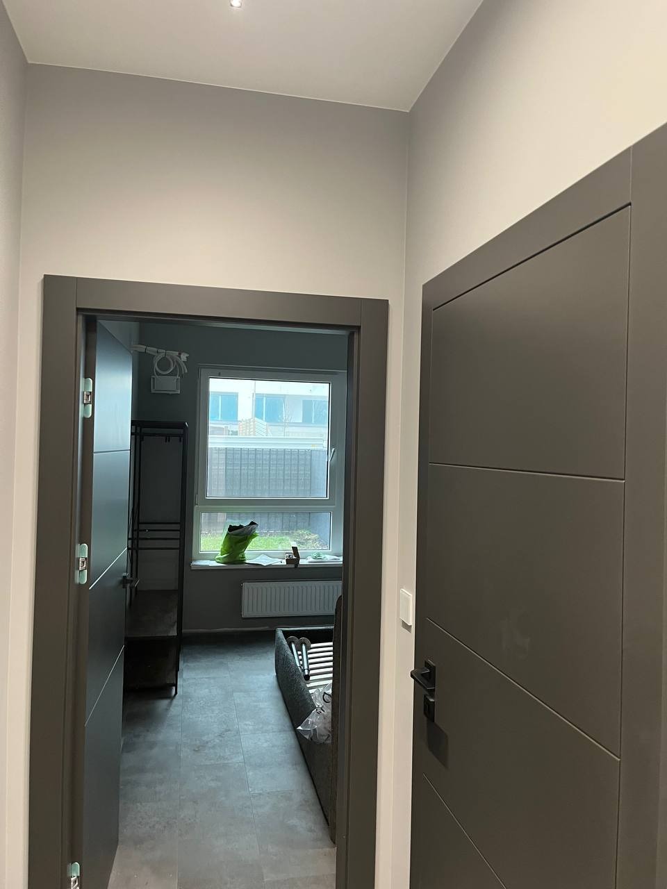 Wnętrze mieszkania po remoncie: świeżo pomalowane ściany w odcieniach szarości, widoczne dwie nowoczesne, grafitowe ościeżnice i drzwi, w tle okno z parapetem i elementami wyposażenia pokoju.