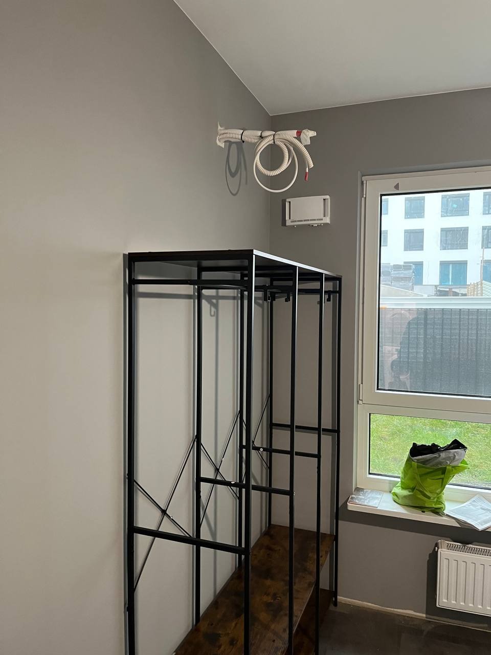 Białe przewody klimatyzacji przymocowane do ściany nad metalowym regałem z drewnianymi półkami, widok na okno z zieloną torbą i dokumentami na parapecie.