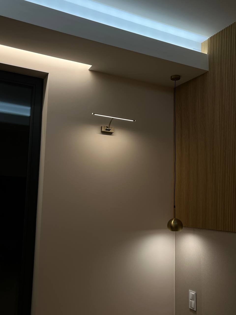 Nowoczesny kinkiet LED nad drzwiami i wisząca lampa kulista z przewodem na tle drewnianej ściany panelowej.