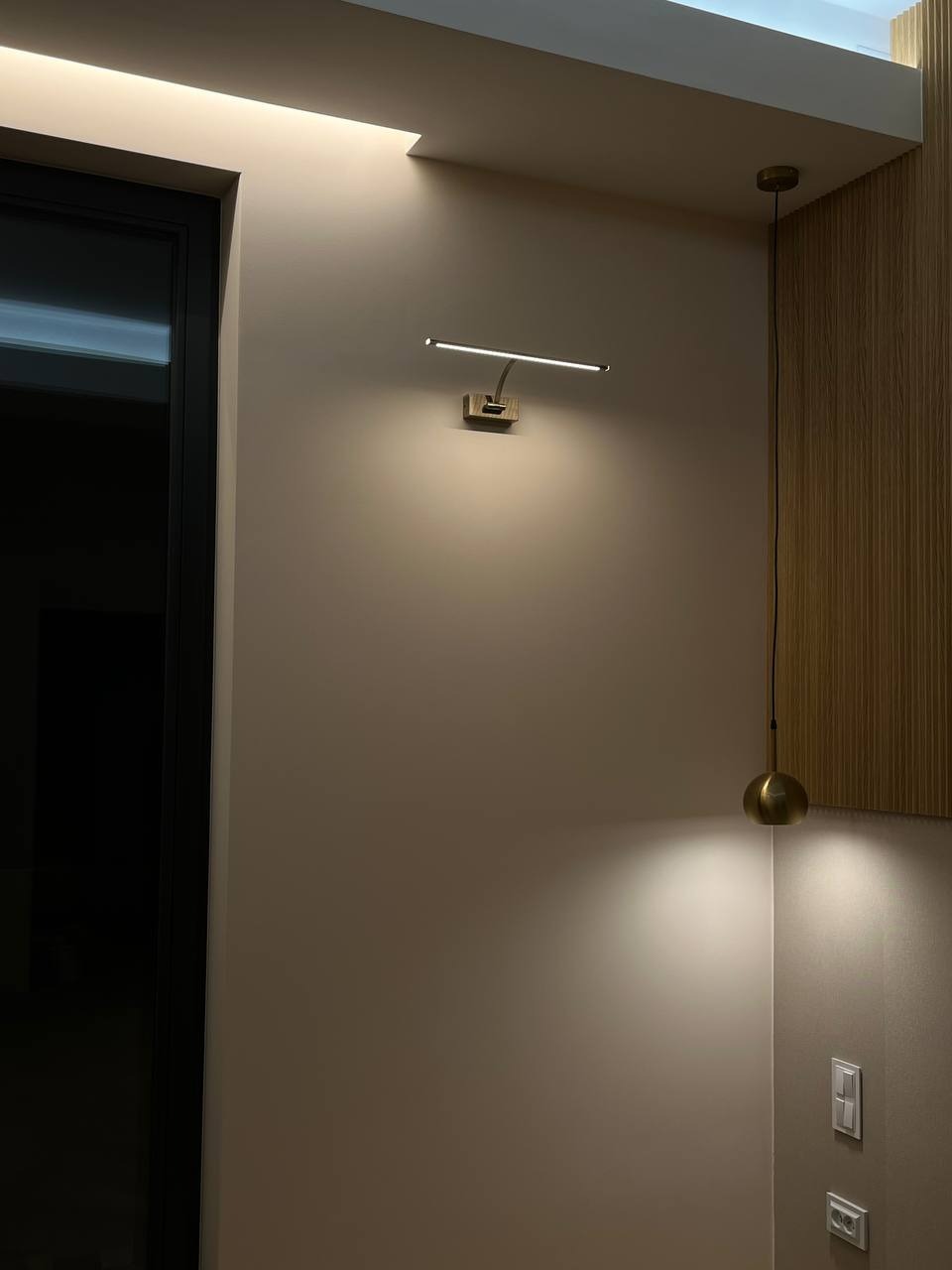 Ściana w odcieniach beżu z zamontowanym nowoczesnym oświetleniem LED w formie listwy, obok wisi lampa wisząca z kulistym kloszem, widoczne gniazdka elektryczne.