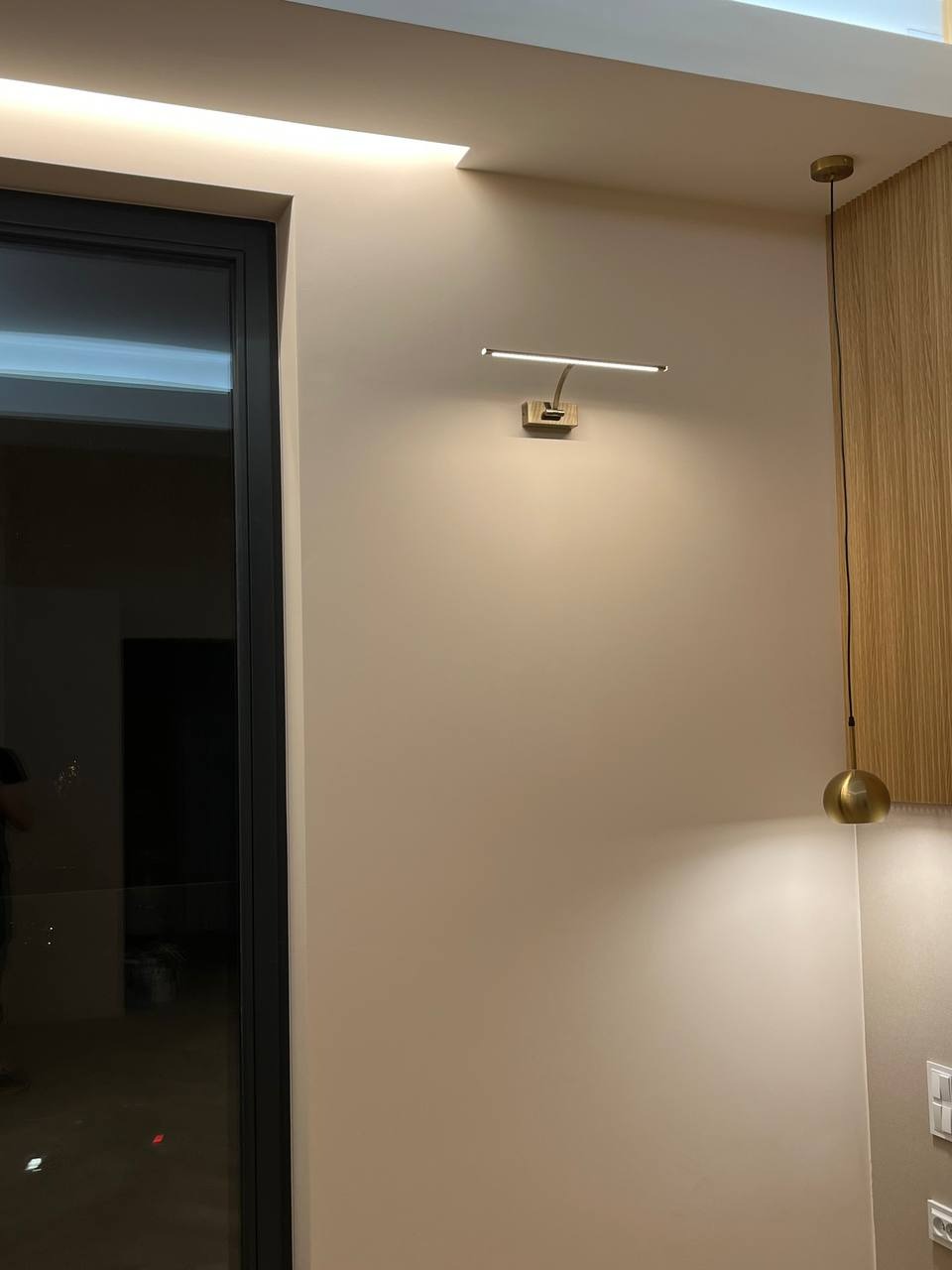 Ściana w kolorze ecru z zamontowanym nowoczesnym oświetleniem ściennym LED z drewnianą podstawą, obok wisi złota lampa wisząca, widoczny fragment drewnianej boazerii.