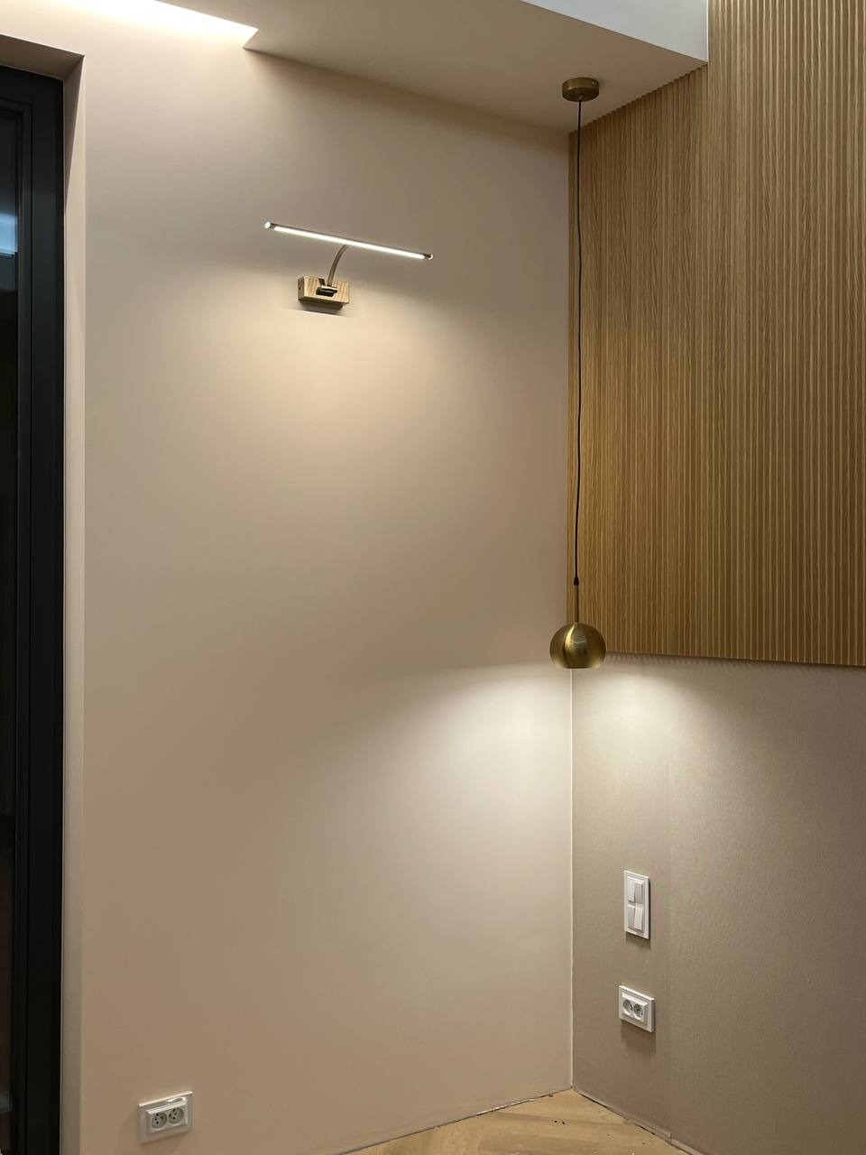 Ściana w jasnym odcieniu z zamontowanym, nowoczesnym oświetleniem: kinkiet LED nad gniazdkiem elektrycznym oraz wisząca lampa kulista z mosiądzu na tle drewnianej boazerii.