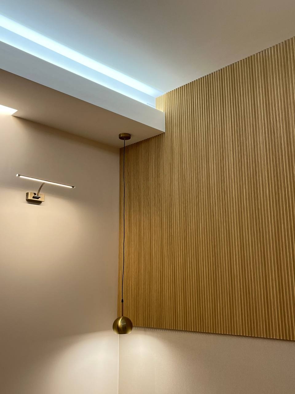 Minimalistyczne wnętrze z oświetleniem: kinkiet nad łóżkiem i wisząca lampa z kulistym kloszem, na tle drewnianej ściany z pionowymi panelami i podświetlanego sufitu.