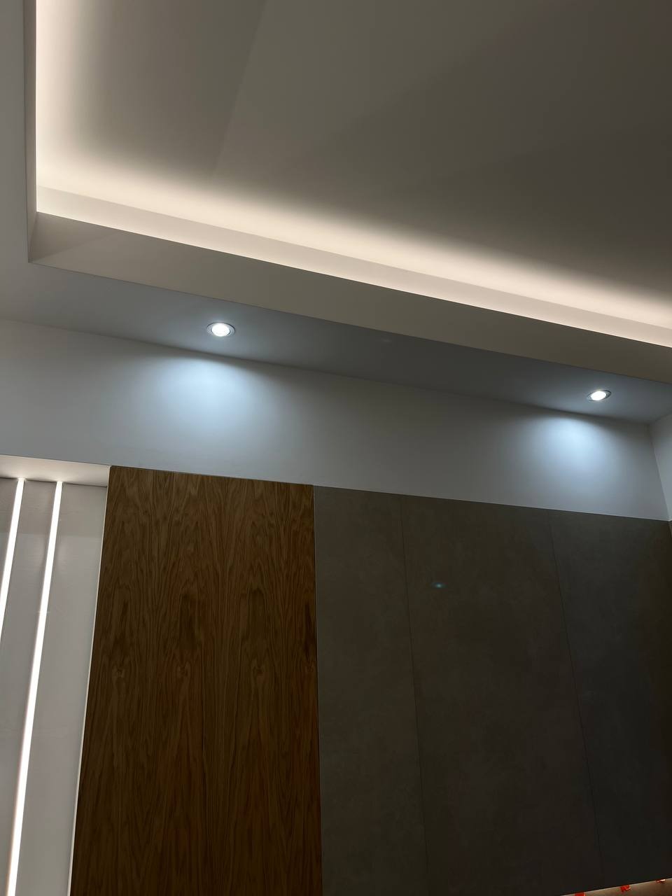 Nowoczesny sufit podwieszany z oświetleniem LED liniowym i punktowym, drewniana okładzina ścienna z pionowymi paskami świetlnymi po lewej stronie i szara ściana po prawej.