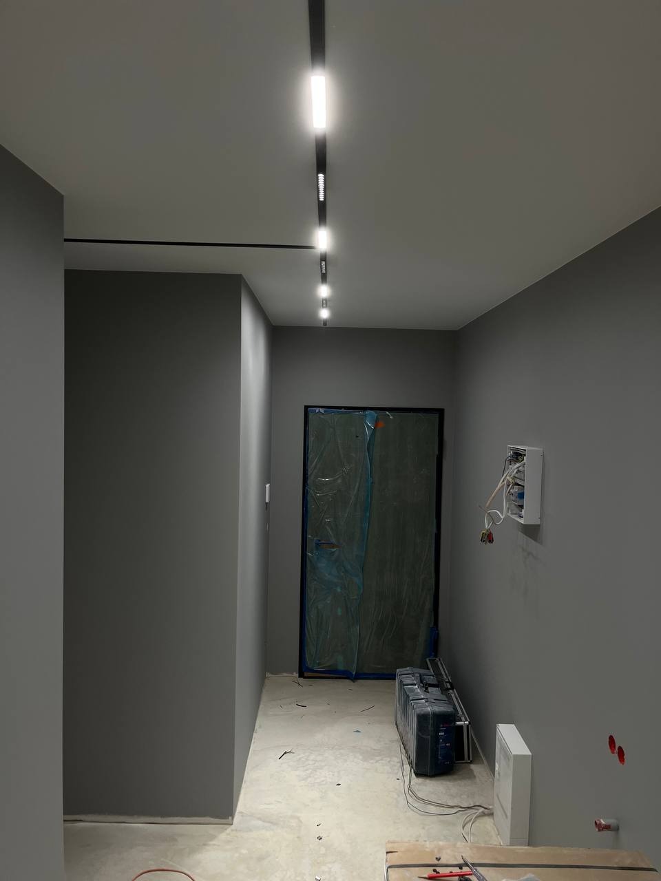 Korytarz w trakcie remontu z zamaskowanymi drzwiami, zamontowanym szynoprzewodem z reflektorami LED i otwartą skrzynką elektryczną na ścianie.