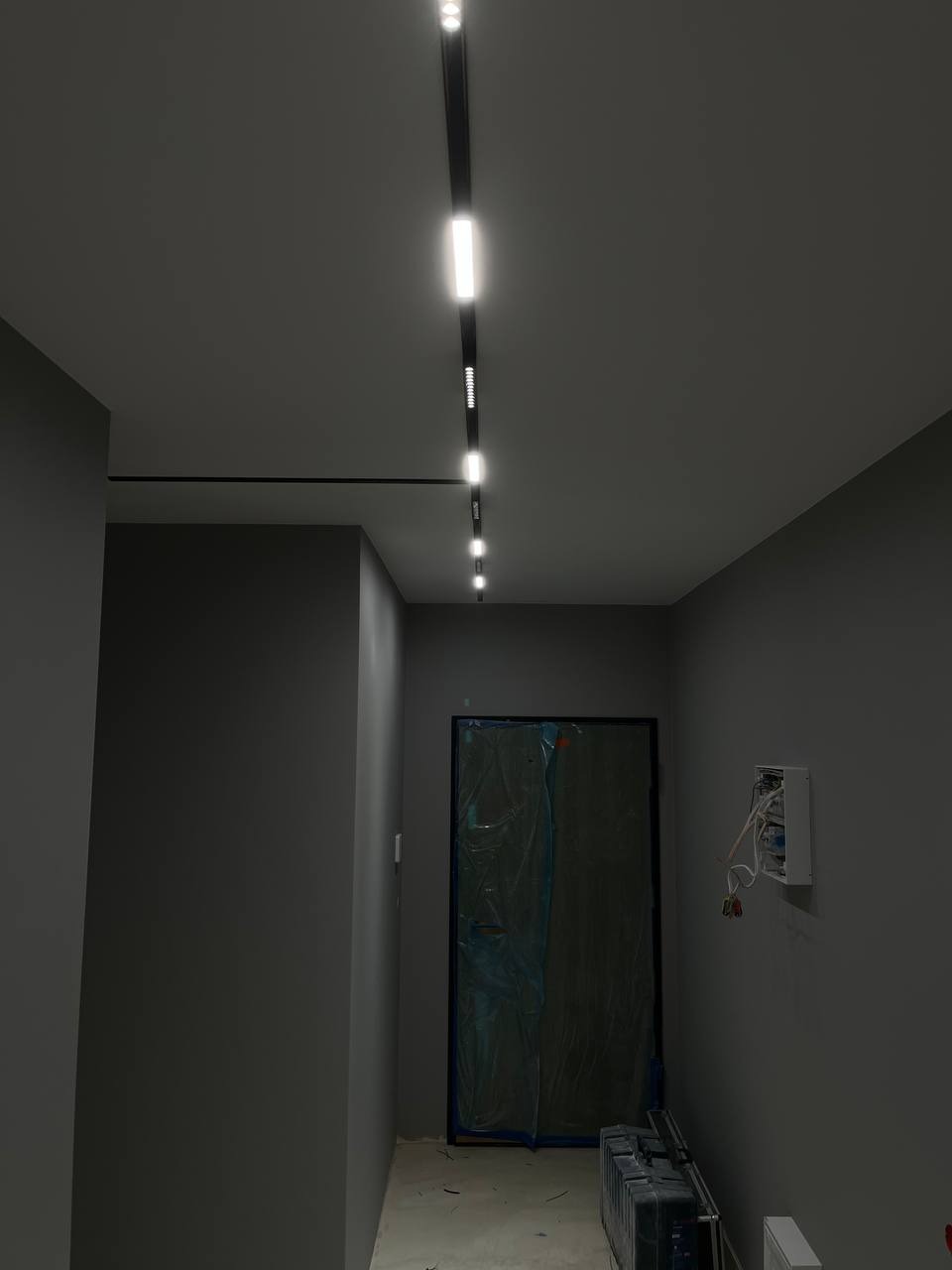 Szary korytarz z zamontowanym systemem szynowym oświetlenia LED, zabezpieczone drzwi folią malarską, skrzynka elektryczna na ścianie.