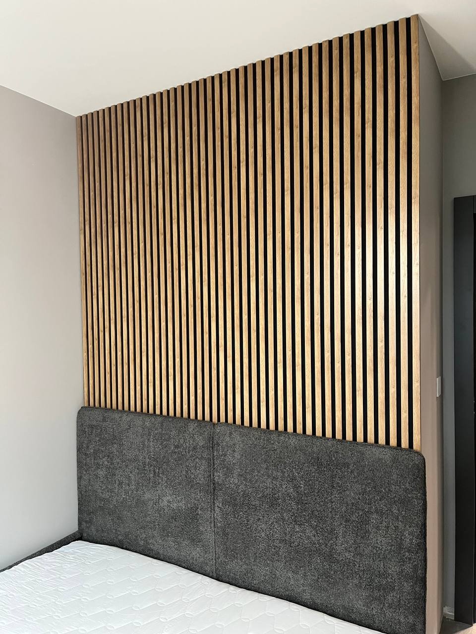 Sypialnia z drewnianą ścianą z pionowych lameli za wezgłowiem łóżka, szara tapicerowana rama łóżka, biały materac.