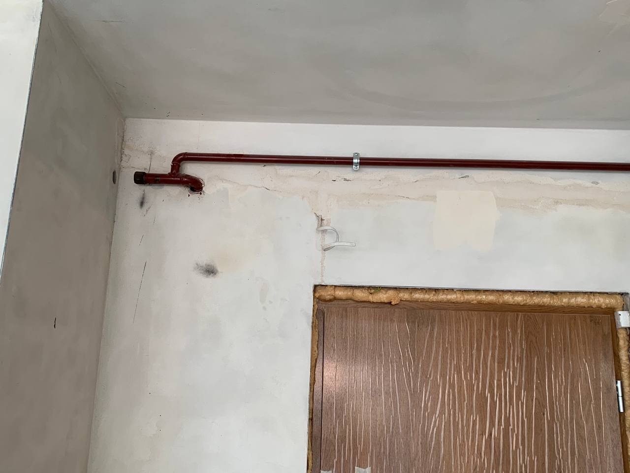 Narożnik pokoju z widoczną rurą grzewczą w kolorze bordowym, przymocowaną do ściany za pomocą uchwytów, nad ościeżnicą drzwiową wypełnioną pianką montażową, ściany częściowo przygotowane do malowania.