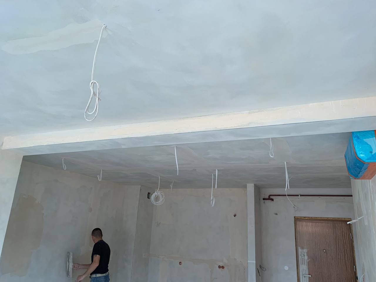 Mężczyzna szpachluje ścianę w pomieszczeniu w stanie deweloperskim, widoczne przewody elektryczne zwisające z sufitu i zabezpieczony klimatyzator.