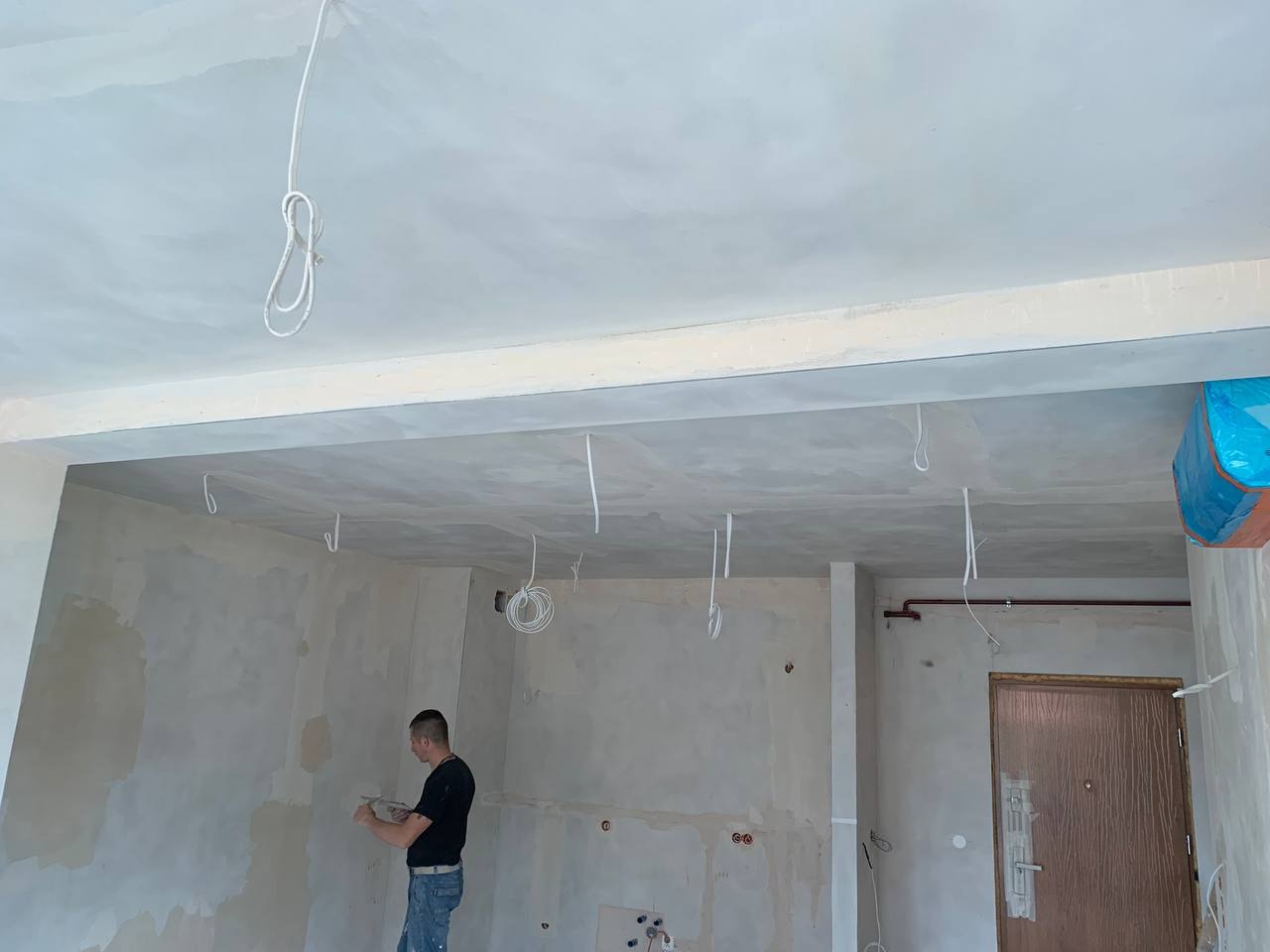 Wnętrze w trakcie remontu: mężczyzna szpachluje ścianę, widoczne wystające przewody elektryczne z sufitu, zakryte drzwi wejściowe.