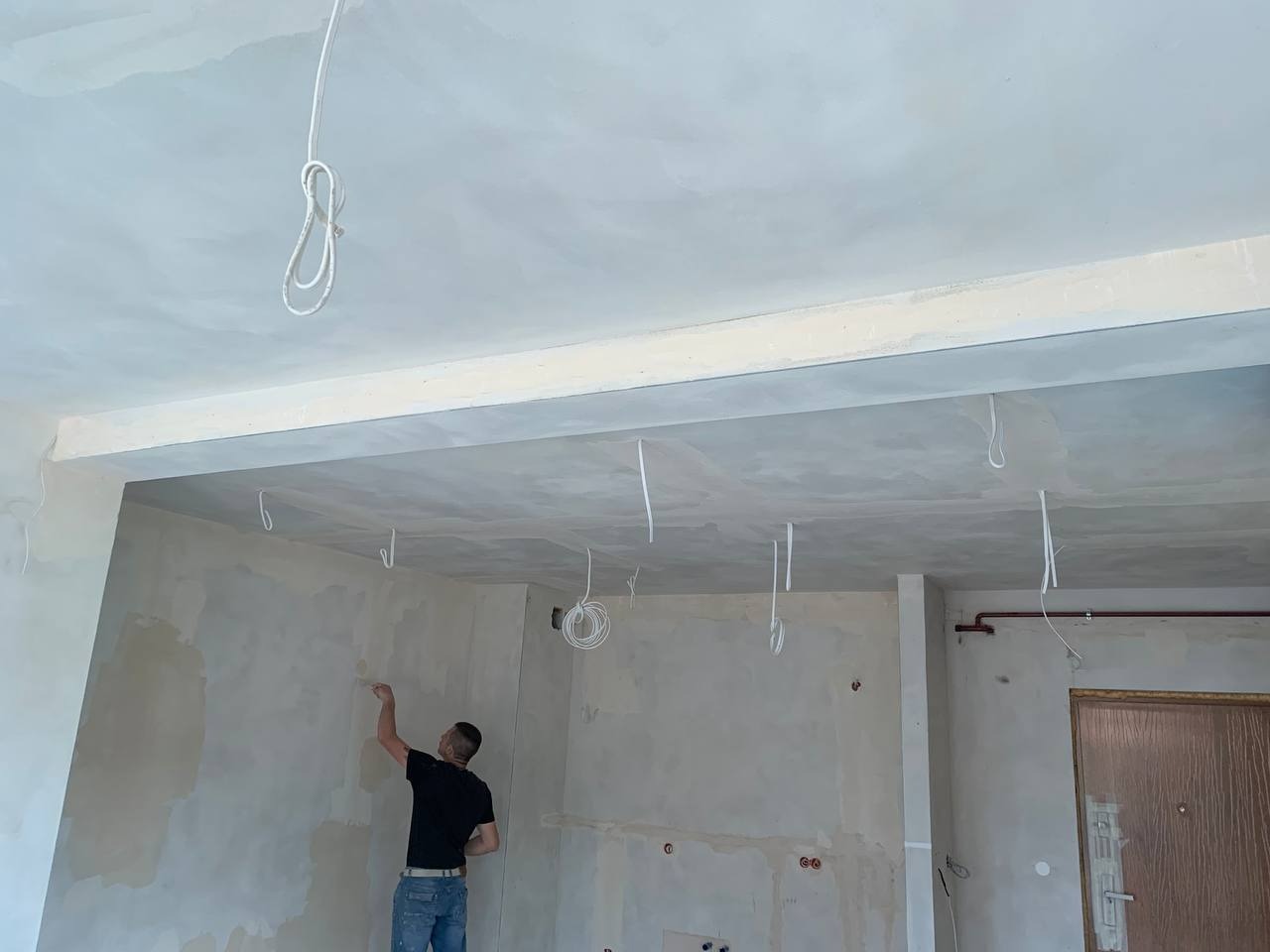 Mężczyzna w czarnej koszulce szpachluje ścianę w surowym wnętrzu z wystającymi przewodami elektrycznymi z sufitu, przygotowując pomieszczenie do malowania.