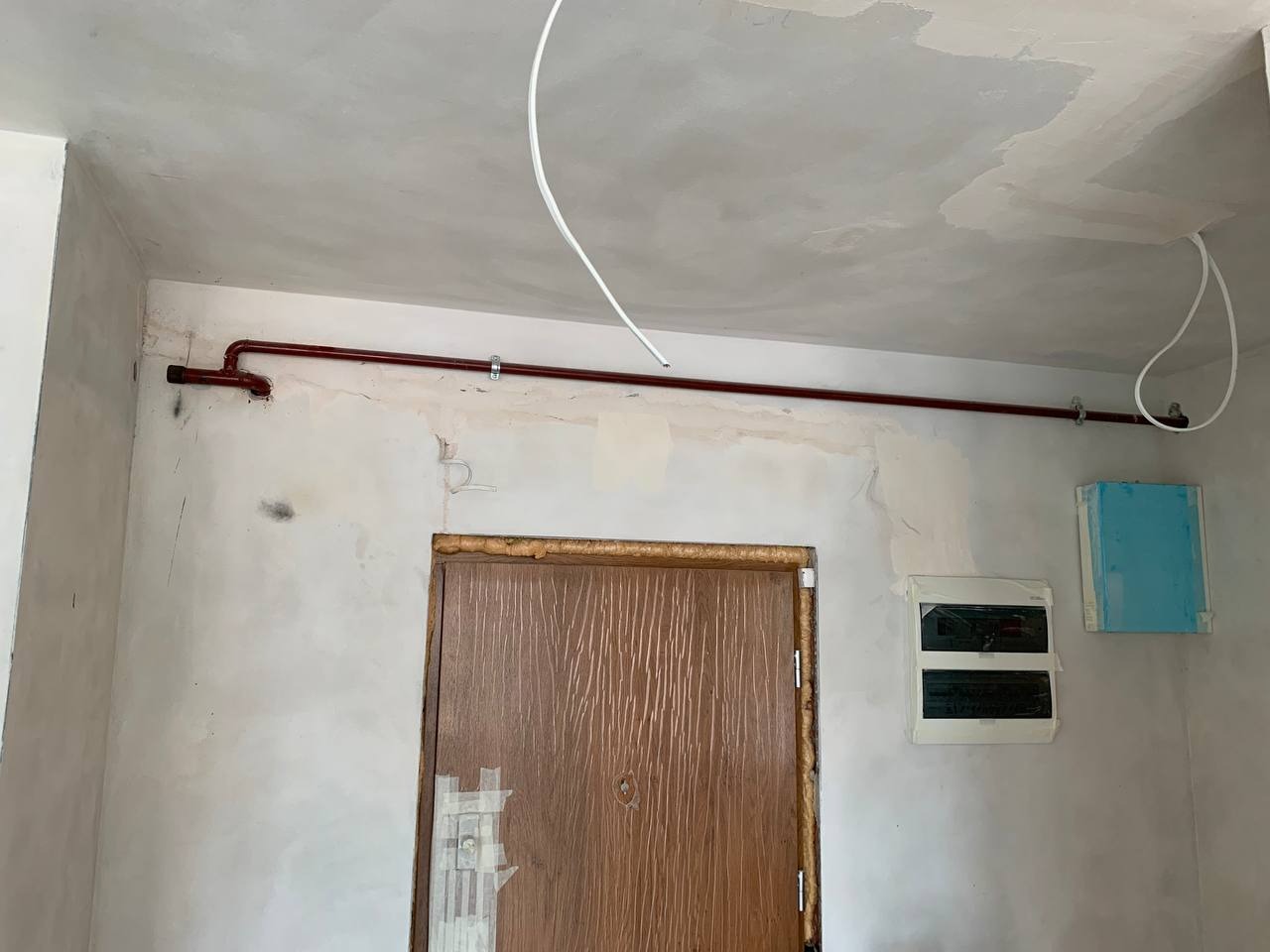Wnętrze w trakcie remontu z widoczną brązową rurą instalacji pod sufitem, niezamaskowaną skrzynką elektryczną i przewodami, ściany w stanie surowym z widocznymi śladami po łataniu i malowaniu.