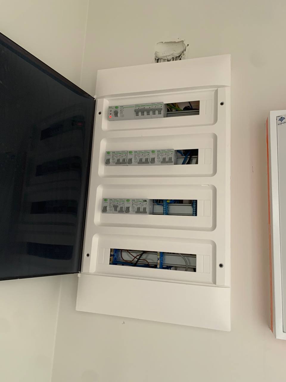 Otwarta biała skrzynka rozdzielcza z widocznymi bezpiecznikami i przewodami, zamontowana na białej ścianie, z widocznym otworem w ścianie nad skrzynką.