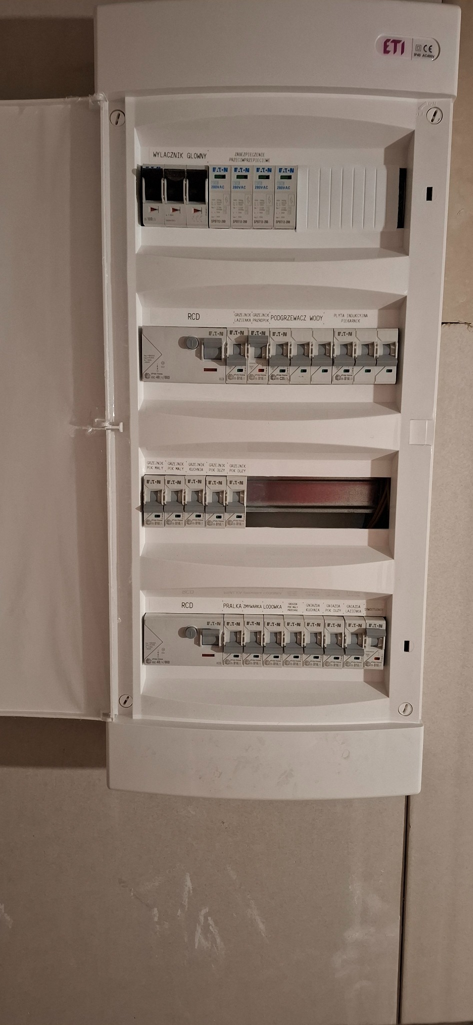 Otwarta skrzynka elektryczna z wyłącznikami EATON, opisana dla różnych obwodów: pralka, zmywarka, lodówka, podgrzewacz wody, płyta indukcyjna. Widoczne zabezpieczenia przeciwprzepięciowe.