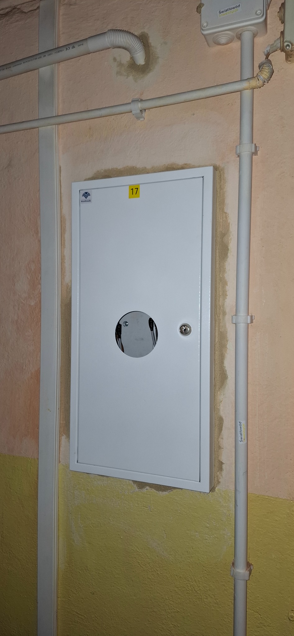 Biała skrzynka z otworem na kable na ścianie z rurami instalacyjnymi. Widoczny numer '17' na żółtej naklejce. Ściana w odcieniach beżu i żółci.