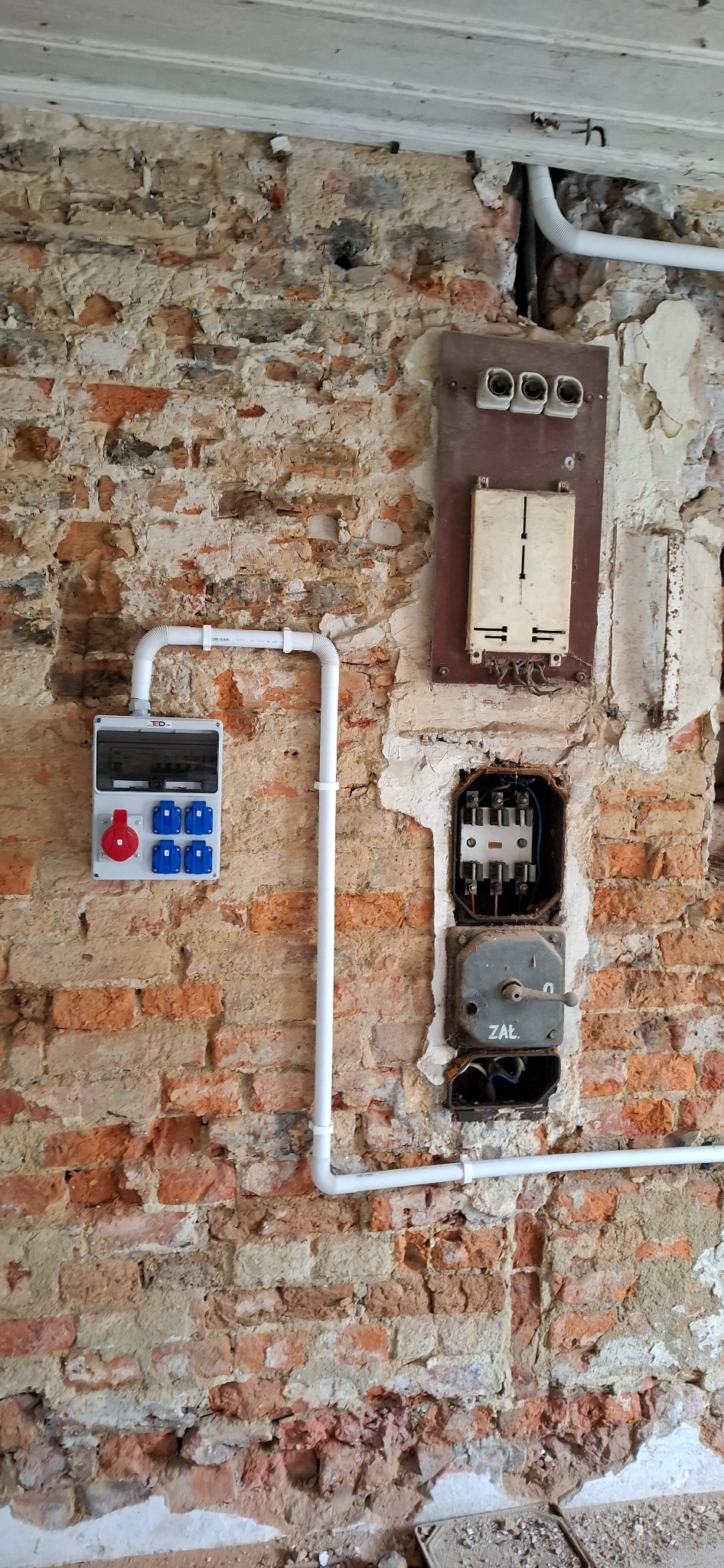 Nowa skrzynka elektryczna z bezpiecznikami i instalacją na tle starej, ceglanej ściany z widocznymi elementami starej instalacji. Kontrast nowego i starego.