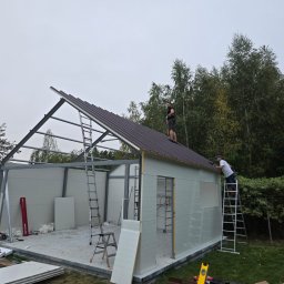 El Toro - Montaż brązowego dachu garażu modułowego z białymi ścianami, dwóch pracowników na dachu i drabinie, widoczne narzędzia i materiały budowlane.