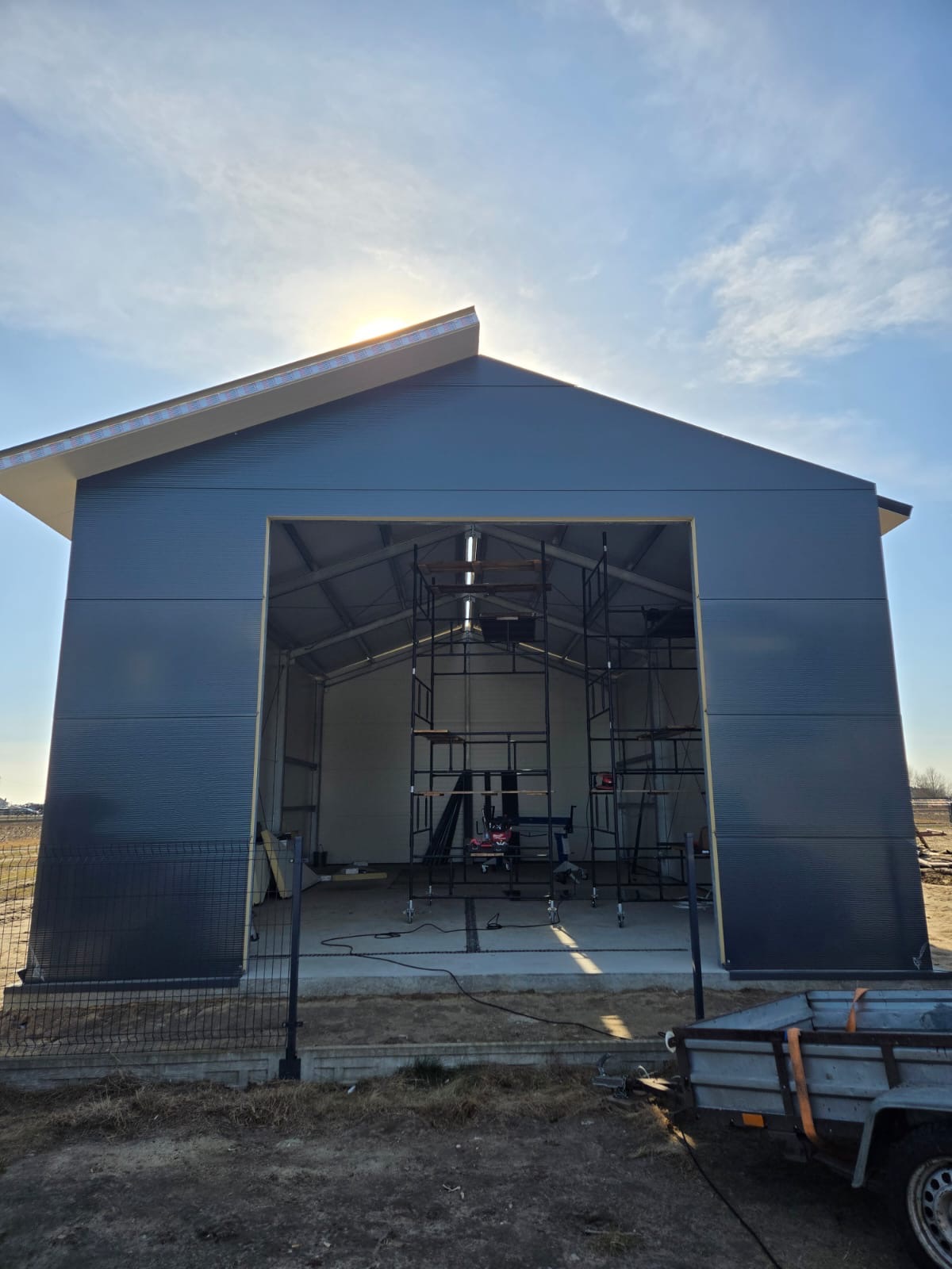 Nowoczesna konstrukcja hali garażowej w trakcie montażu, widoczne rusztowania wewnątrz, niebieska blacha trapezowa na elewacji, w tle błękitne niebo z promieniami słońca.