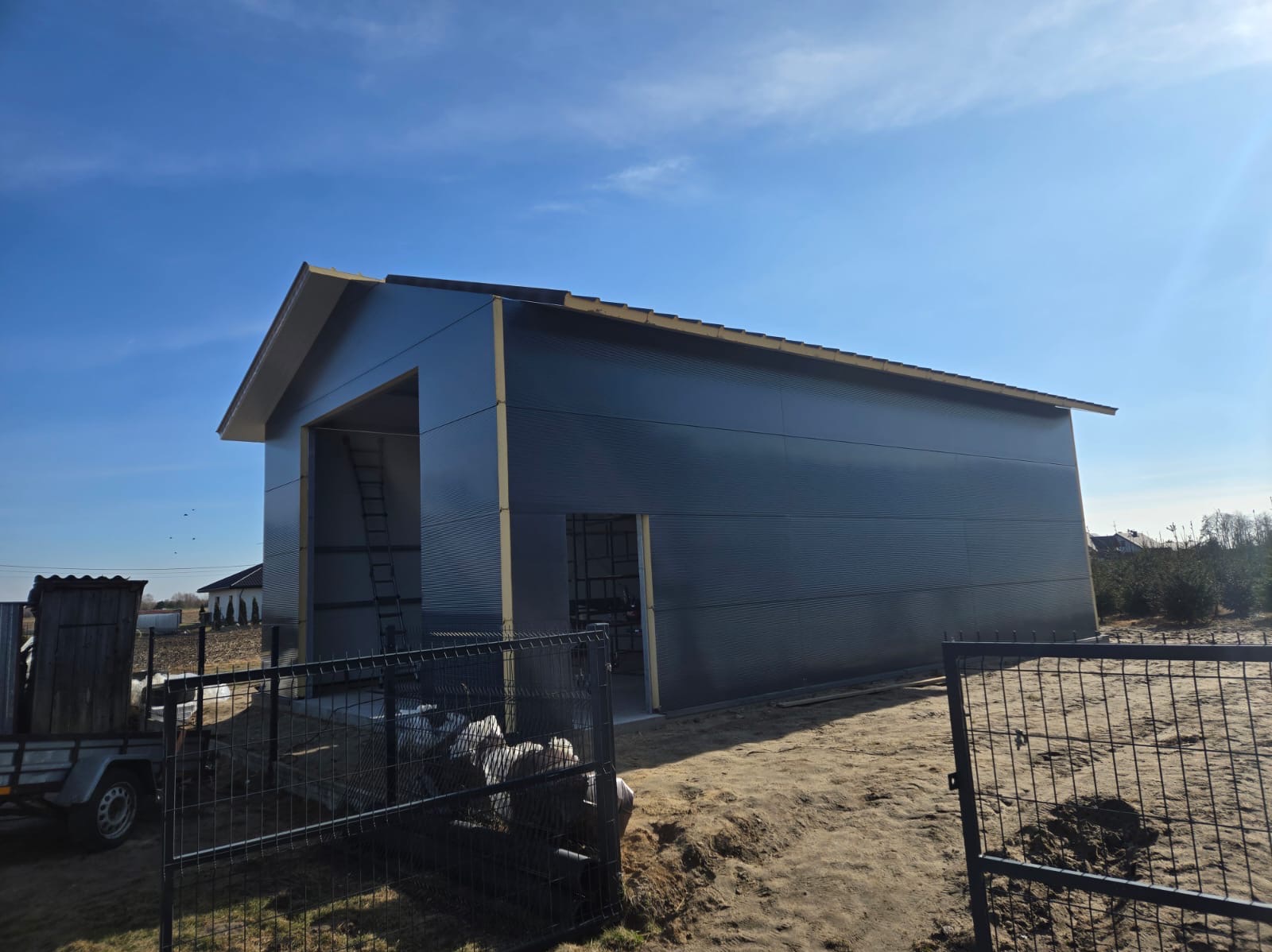 Nowoczesna, metalowa konstrukcja w kolorze grafitowym, prawdopodobnie magazynowa, z widocznymi drzwiami i otwartą przestrzenią wjazdową, częściowo ogrodzona czarnym płotem z siatki, na tle...