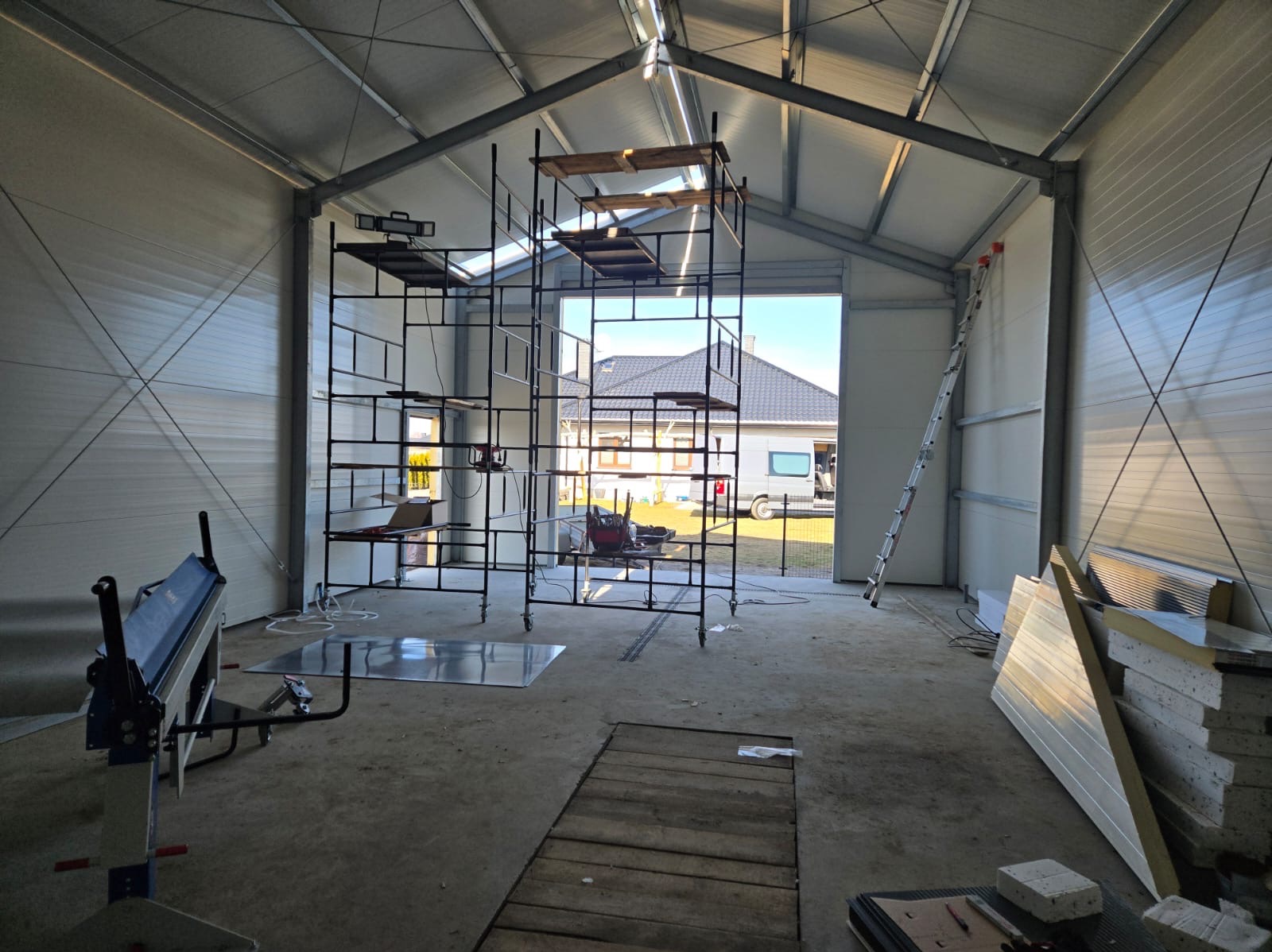 Wnętrze garażu w trakcie budowy lub remontu, widoczne rusztowanie, blacha, narzędzia i drabina, z otwartym wjazdem na zewnątrz.