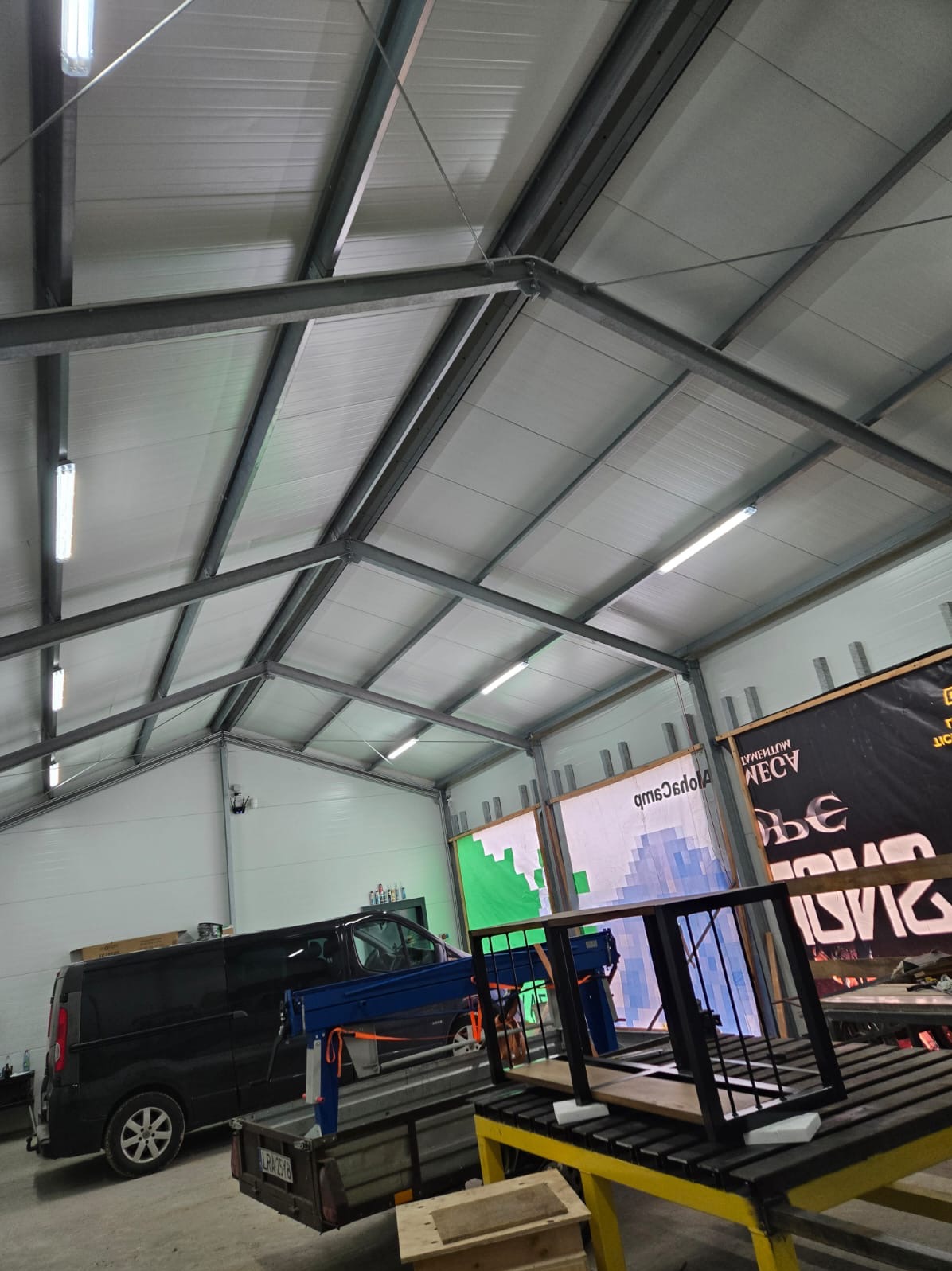 Warsztat z konstrukcją metalową, prawdopodobnie element bramy, na żółtym stole roboczym, widoczny bus i przyczepa w tle, oświetlenie sufitowe.