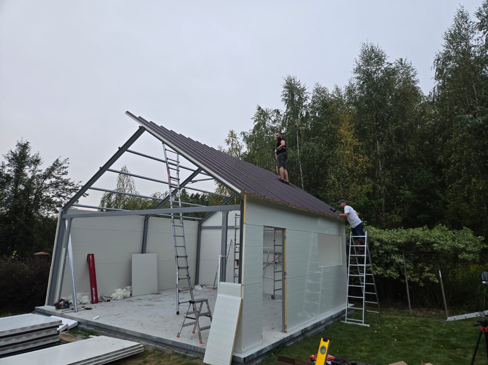 Montaż brązowego dachu garażu modułowego z białymi ścianami, dwóch pracowników na dachu i drabinie, widoczne narzędzia i materiały budowlane.