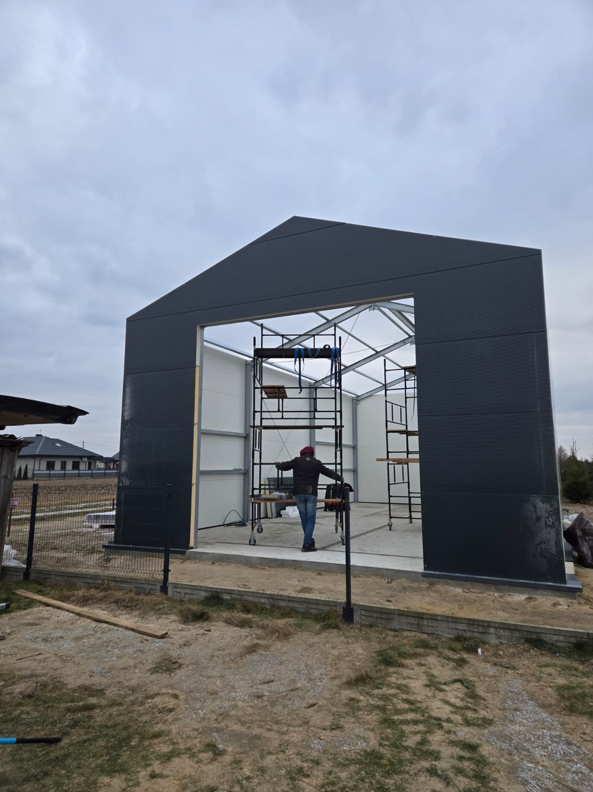 Montaż szarej konstrukcji garażowej z blachy trapezowej, widoczne rusztowania i pracownik wewnątrz, w tle zabudowa jednorodzinna.
